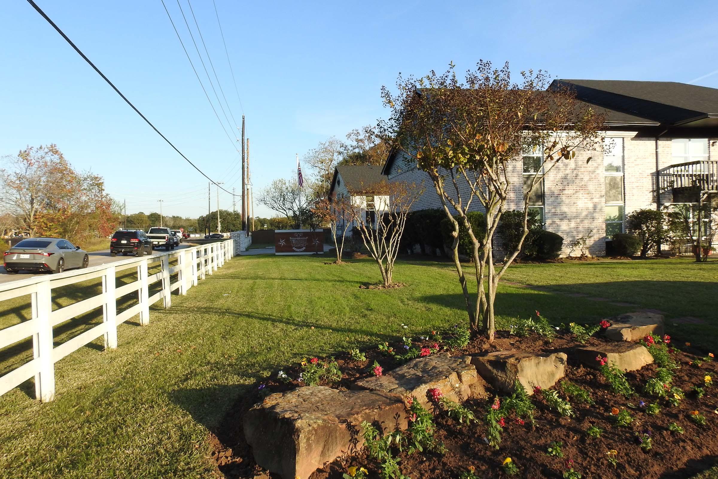 A well-maintained grassy area featuring flower beds with colorful blooms, a small decorative rock formation, and trees lining the front. A white fence runs alongside the street, with cars parked nearby and a building visible in the background against a clear blue sky.