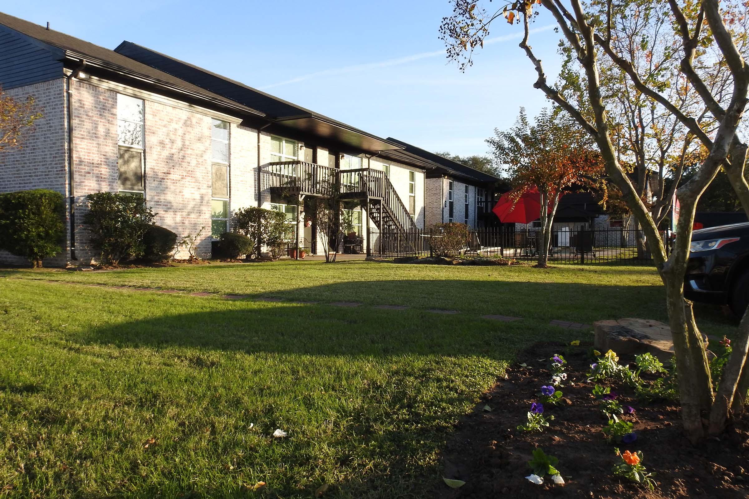 A multi-unit apartment building with a wooden staircase leading to upper floors, surrounded by well-maintained grass and small flower beds. There are trees and a red canopy visible in the background, with a partially fenced yard. The lighting suggests late afternoon or early evening.