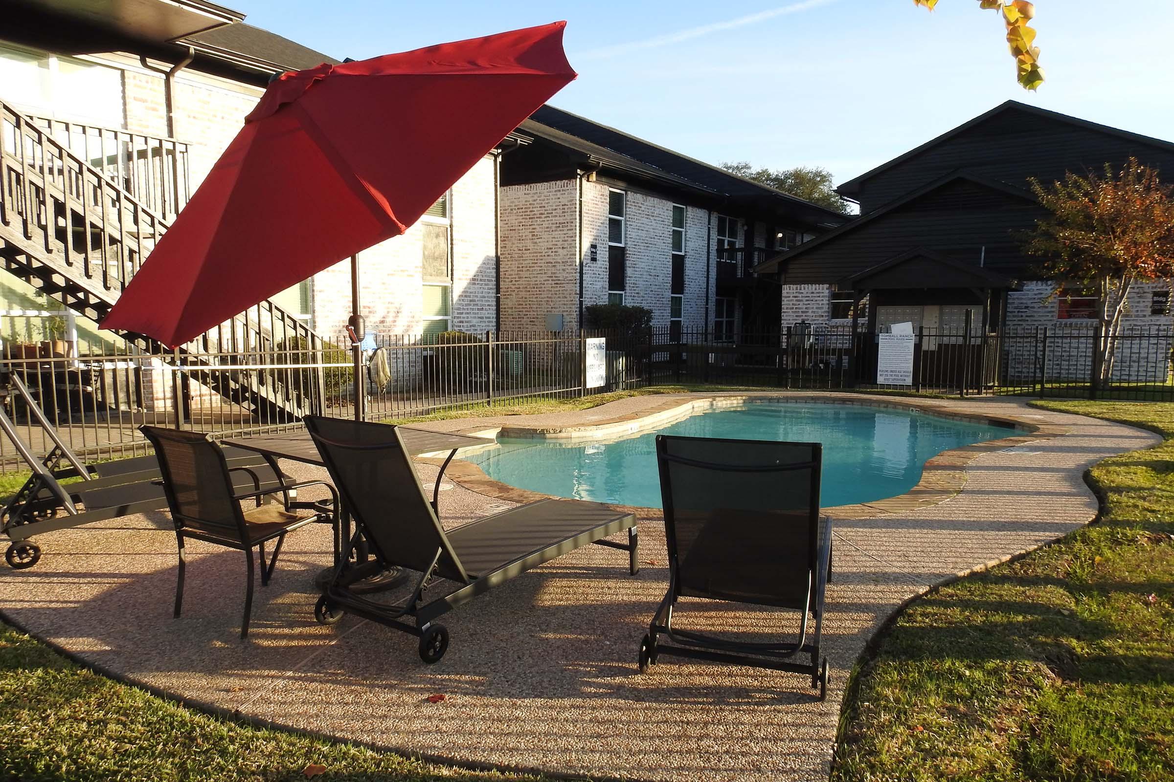 A pool area featuring a small swimming pool surrounded by a concrete deck. Two lounge chairs are positioned nearby, with one chair being partially shaded by a large red umbrella. In the background, there are two buildings with a mix of brick and dark siding, along with green grass and a clear sky.