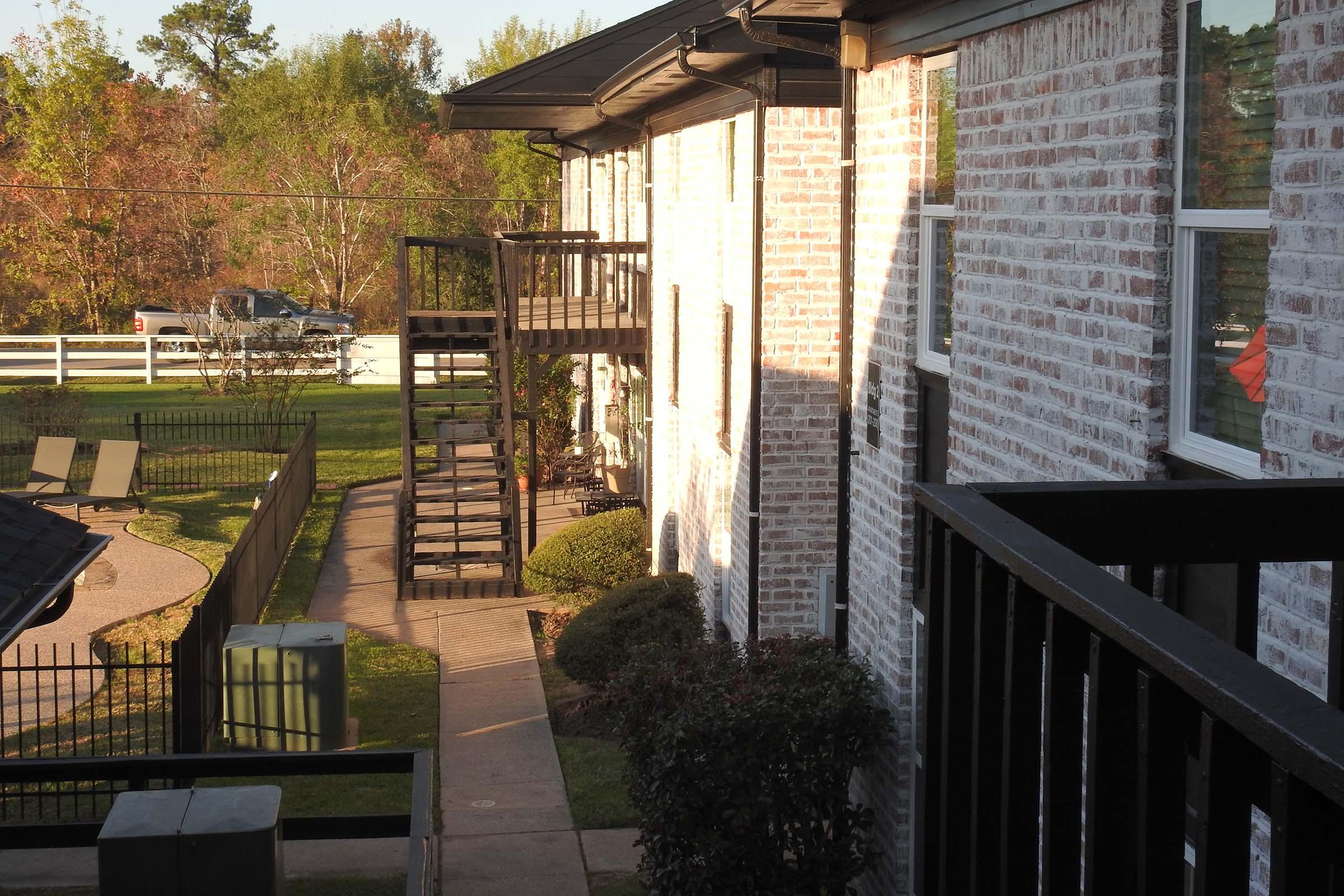 A view of a brick apartment building featuring a pathway, stairs, and greenery. The scene includes a fenced area, a truck parked nearby, and trees with autumn foliage in the background. The lighting suggests late afternoon or early evening.