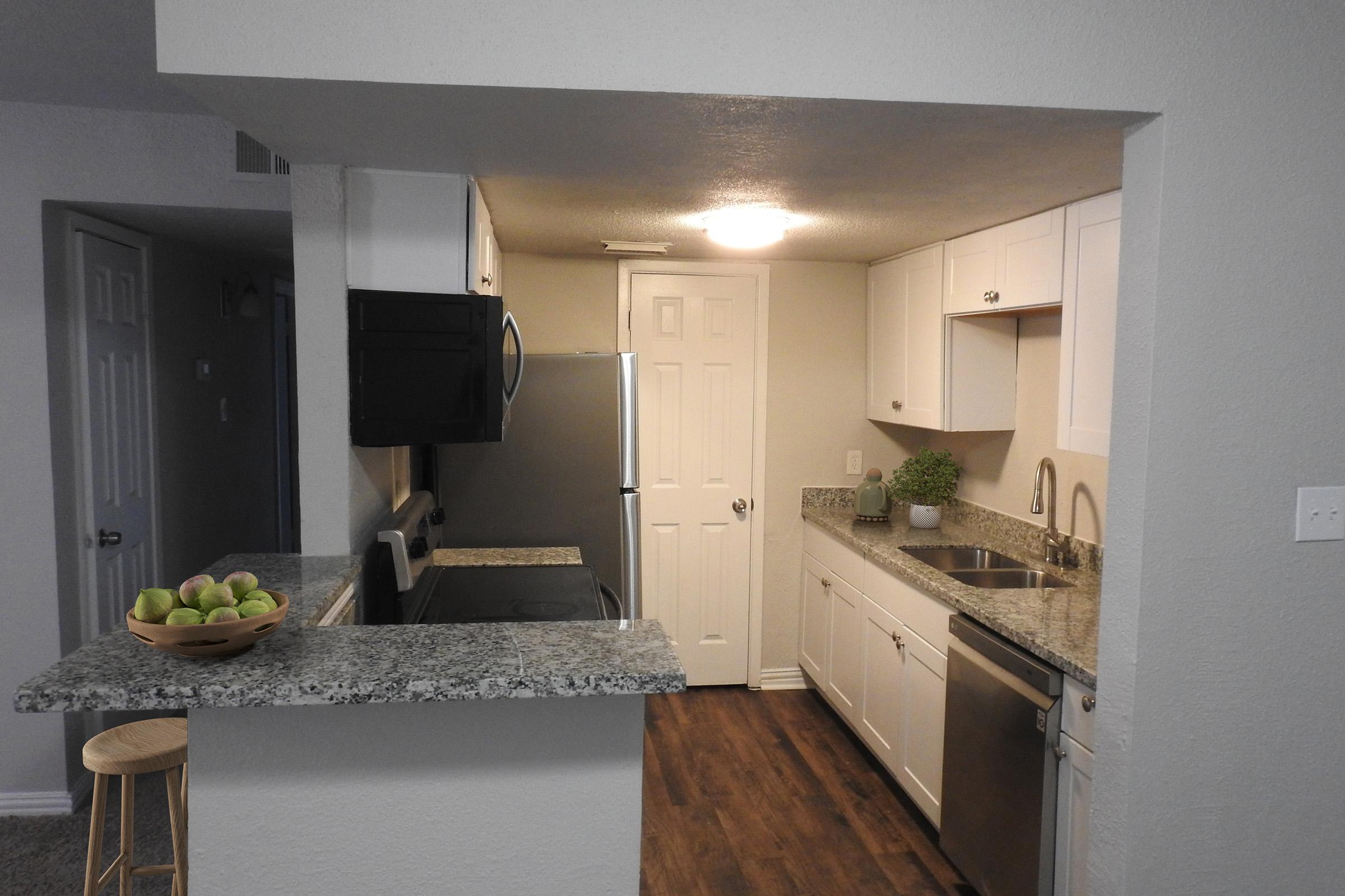 A modern kitchen with white cabinetry and granite countertops. Features include a stainless steel refrigerator, oven, and sink. There's a small bar area with a stool and a bowl of green apples. The walls are light-colored, and the flooring is a wood-like laminate. Bright lighting enhances the open space.