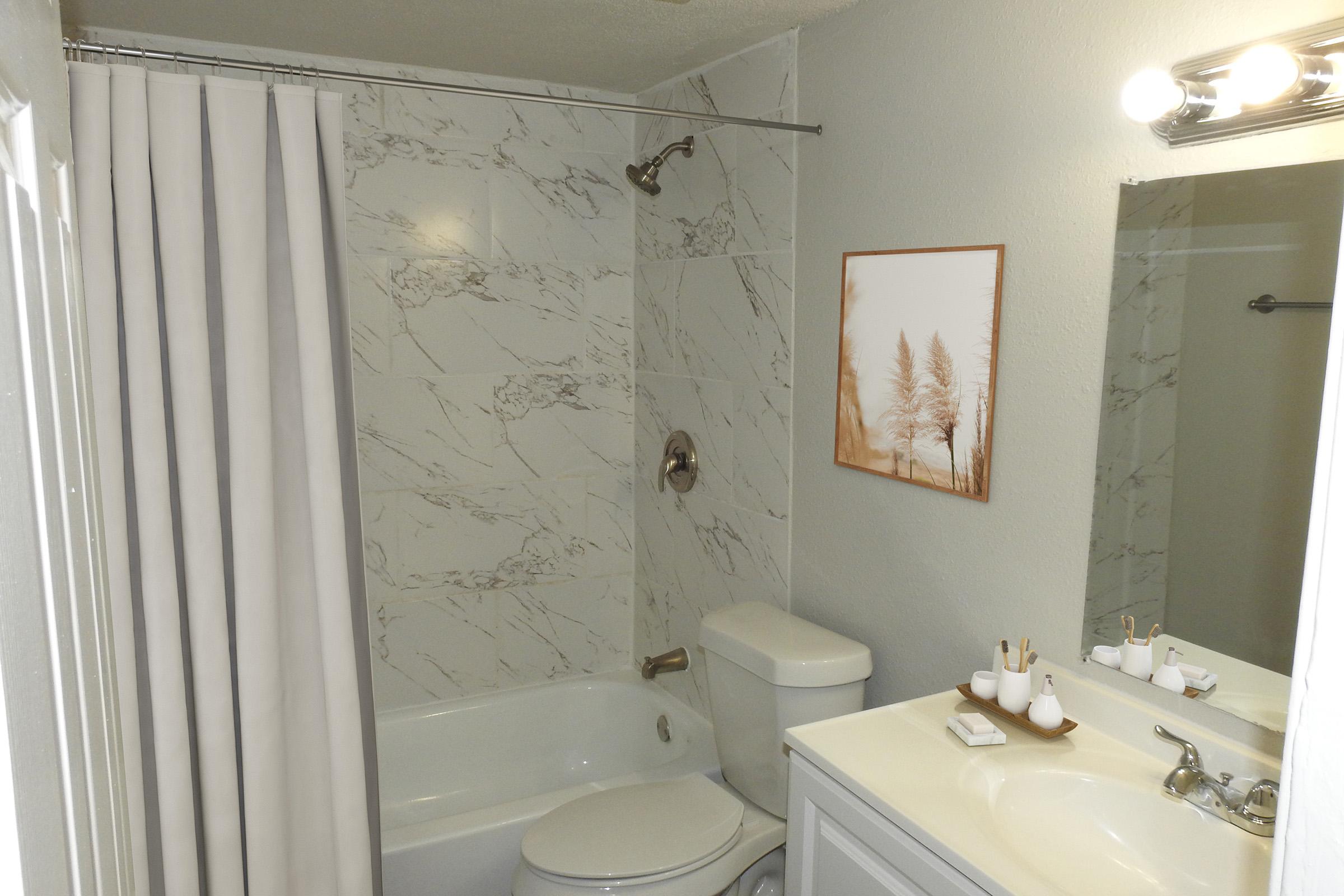 A clean, modern bathroom featuring a white tub, shower with marble-like tiles, a white toilet, and a vanity with a sink. A simple artwork of trees adorns the wall, and a shower curtain hangs beside the tub. The space is well-lit and organized with toiletries neatly arranged on the counter.