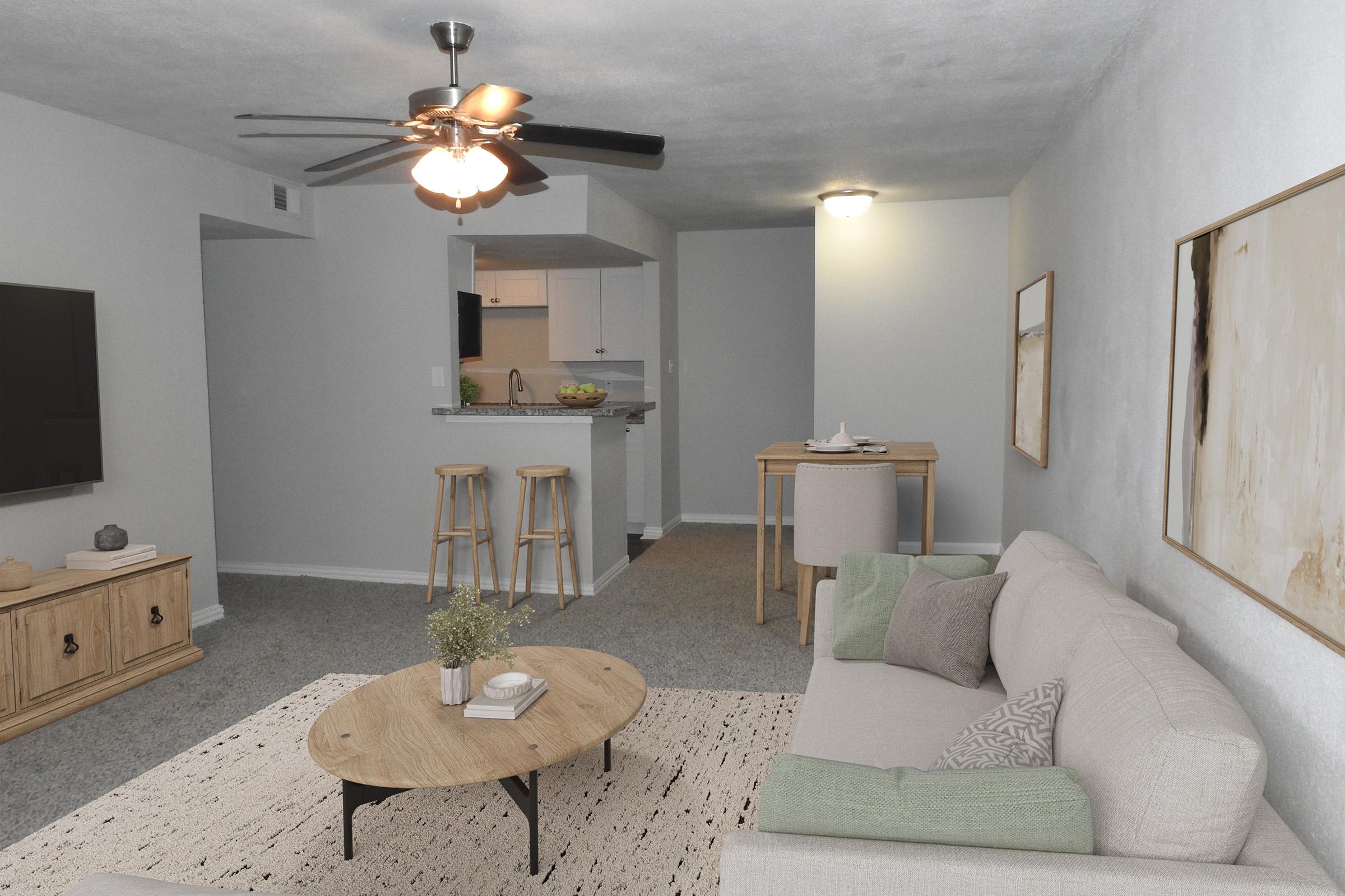 A cozy living room featuring a light-colored sofa with decorative pillows, a round coffee table, a ceiling fan, and a TV mounted on the wall. To the right, there's a small dining area with a table and stools. The space has neutral walls and a soft carpet, creating a welcoming atmosphere.