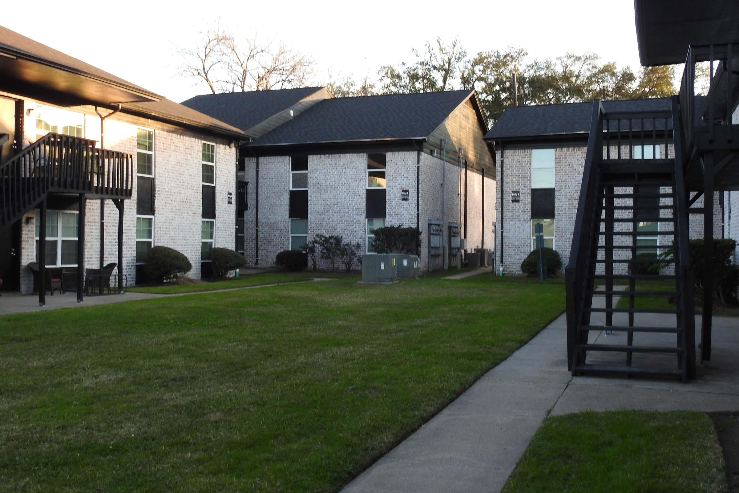 View of an apartment complex courtyard featuring two multi-story buildings with white brick exteriors. The scene includes a green lawn, shrubs, and a staircase leading to a second-floor entrance. The sky is clear, suggesting an afternoon setting.