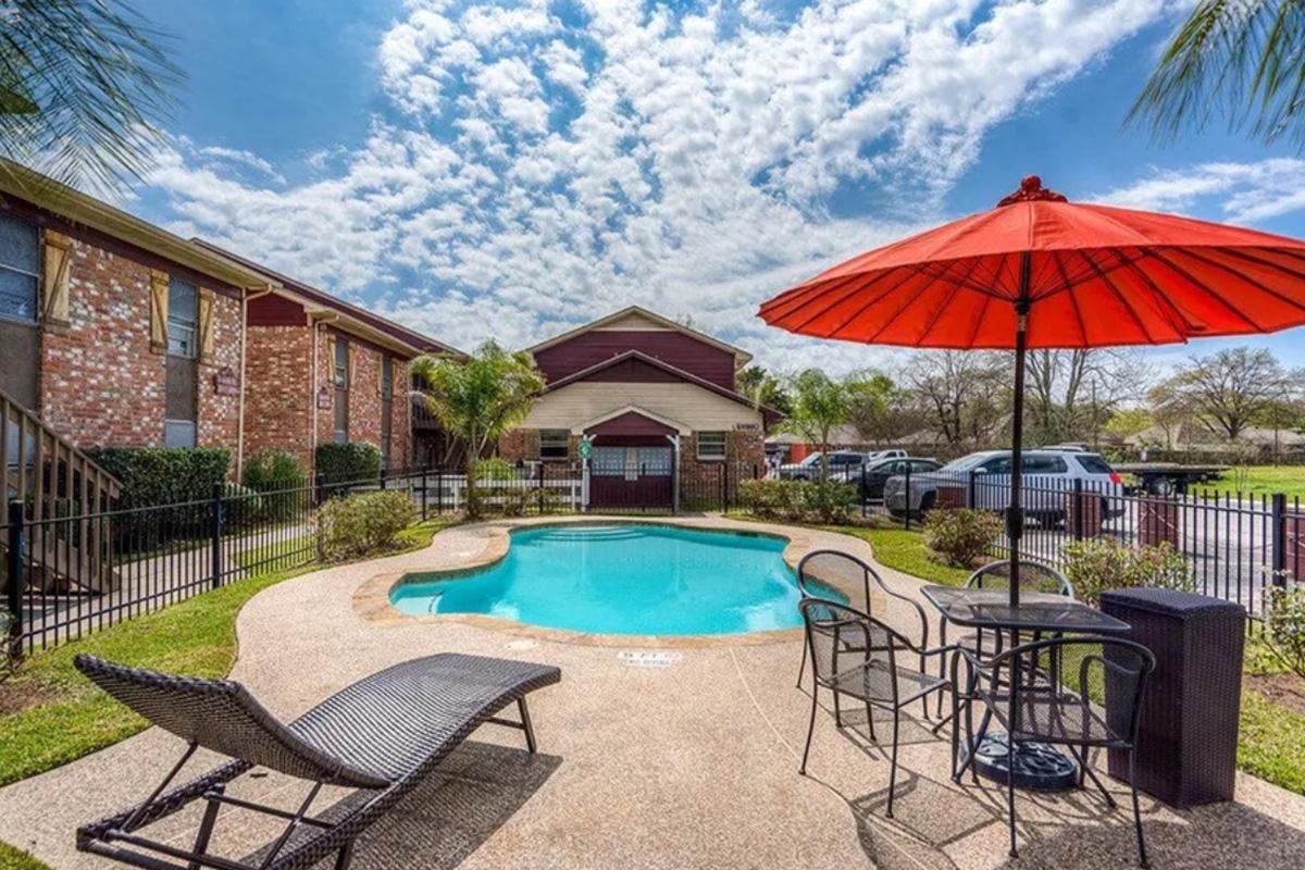 A serene outdoor pool area featuring a circular pool surrounded by lounge chairs and a vibrant red umbrella. Lush green landscaping and a fence create a private atmosphere, with brick buildings visible in the background and a clear blue sky scattered with fluffy clouds.