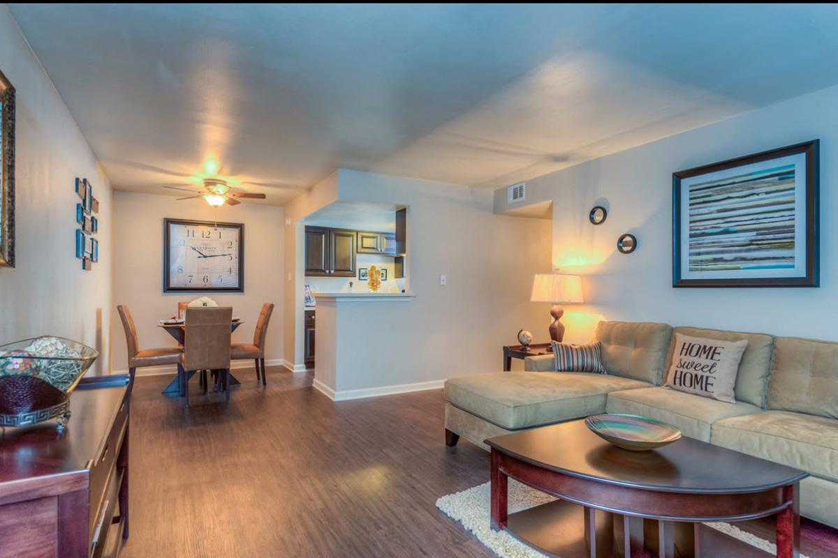 A cozy living room featuring a light brown sectional sofa, a round coffee table, and decorative cushions. The space is well-lit with a ceiling fan, showcasing a dining area in the background with a table and chairs. The walls are adorned with modern artwork and a clock.