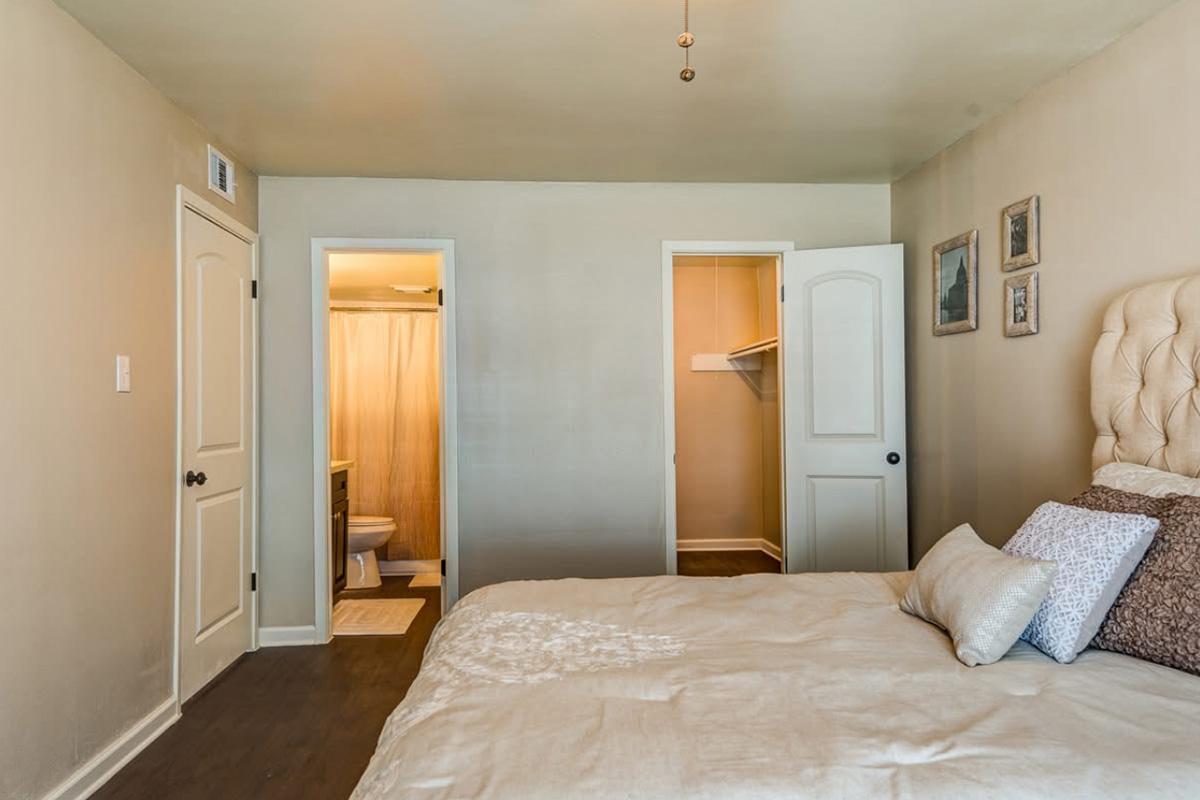 A cozy bedroom featuring a queen-sized bed with decorative pillows. The room has light-colored walls and a smooth wood floor. Two doors lead to a small bathroom and a closet. Natural light filters in, creating a warm atmosphere. Framed pictures adorn the wall, adding a personal touch.