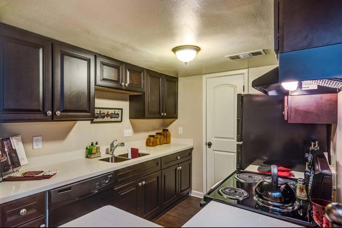 A modern kitchen featuring dark wood cabinets, a white countertop, and stainless steel appliances. It includes a black stove, a sink, and a door leading to another room. Decorative items like jars and a framed picture are arranged on the countertop. Soft lighting creates a warm ambiance.