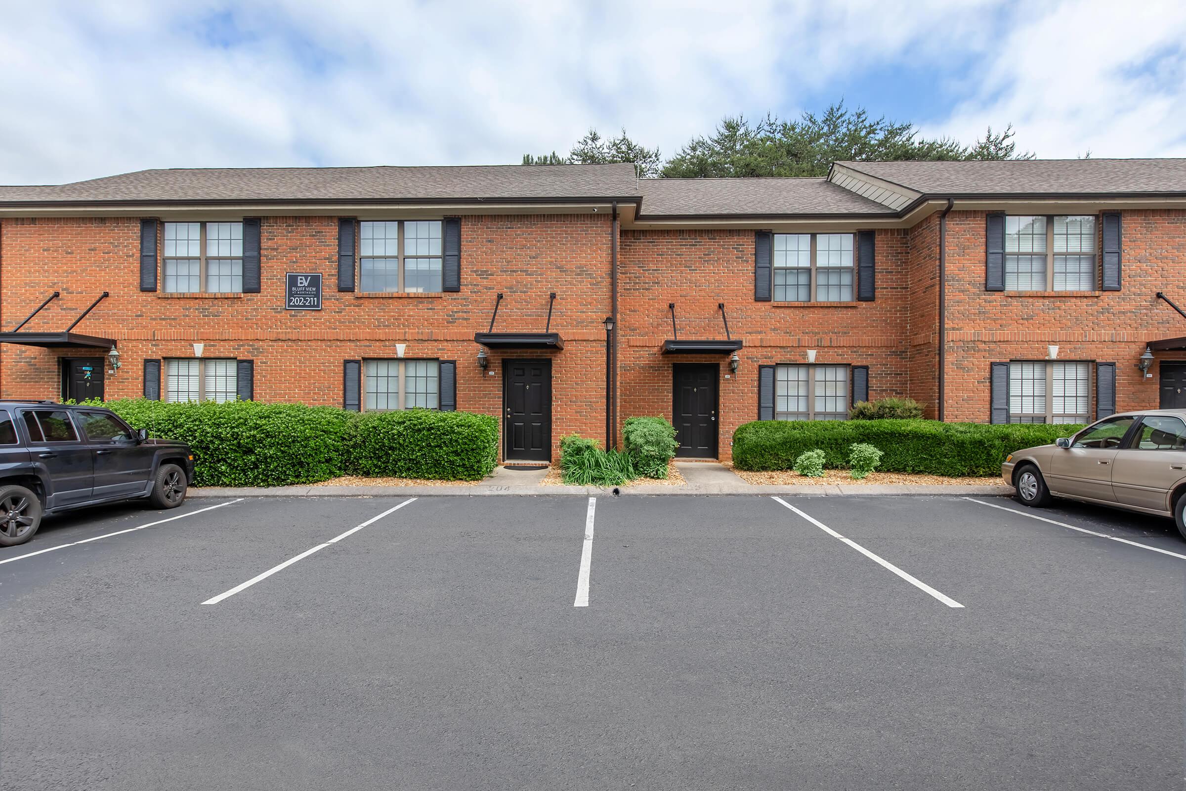 A row of two-story brick apartment units with black doors. The building is flanked by low greenery and a parking lot with several parked cars. The sky is partly cloudy, adding to the tranquil suburban atmosphere.