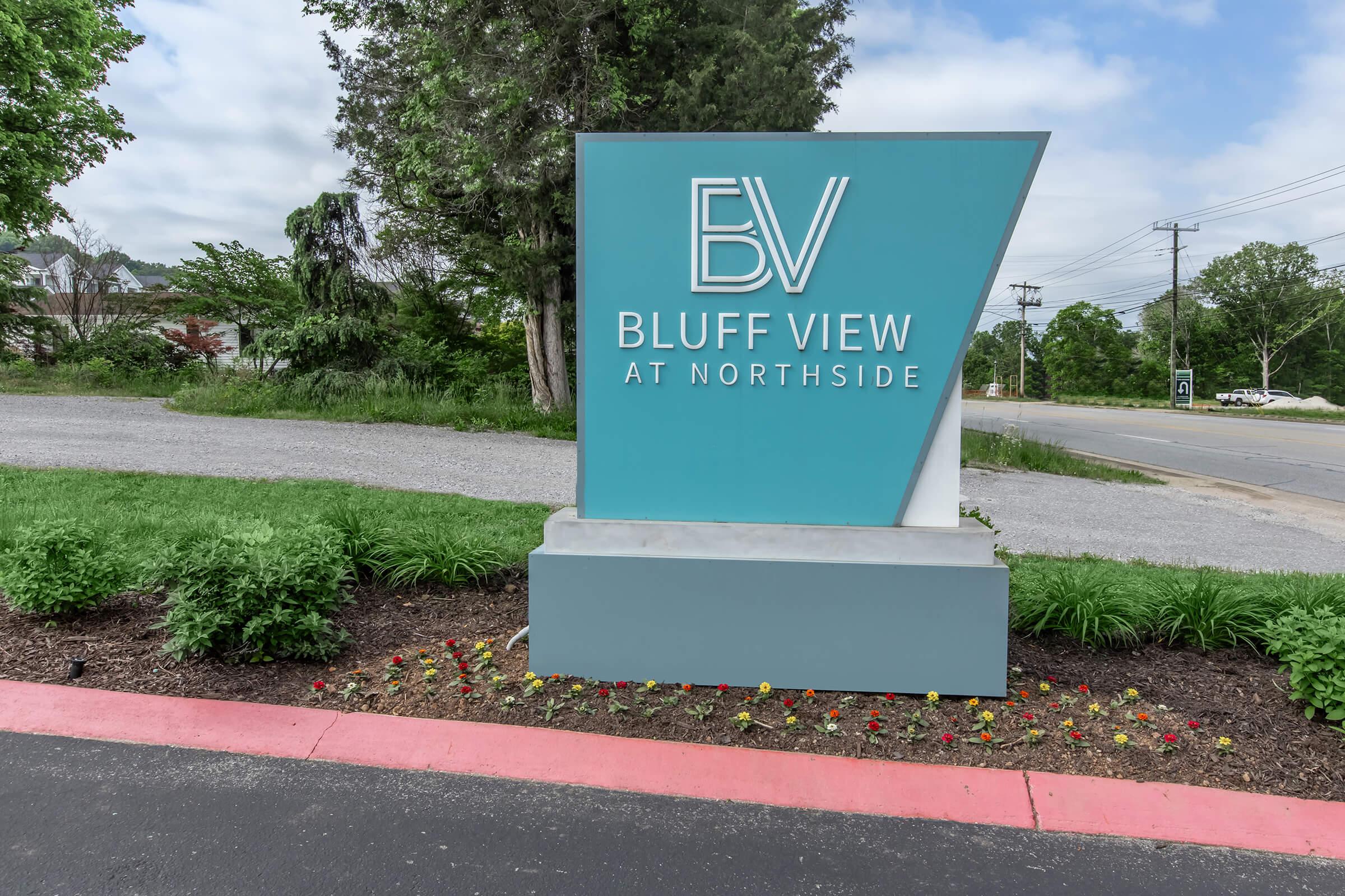 Sign for Bluff View at Northside, featuring a modern design with teal and gray colors. Surrounded by greenery and flowers, the sign is set along a road with a cloudy sky in the background.