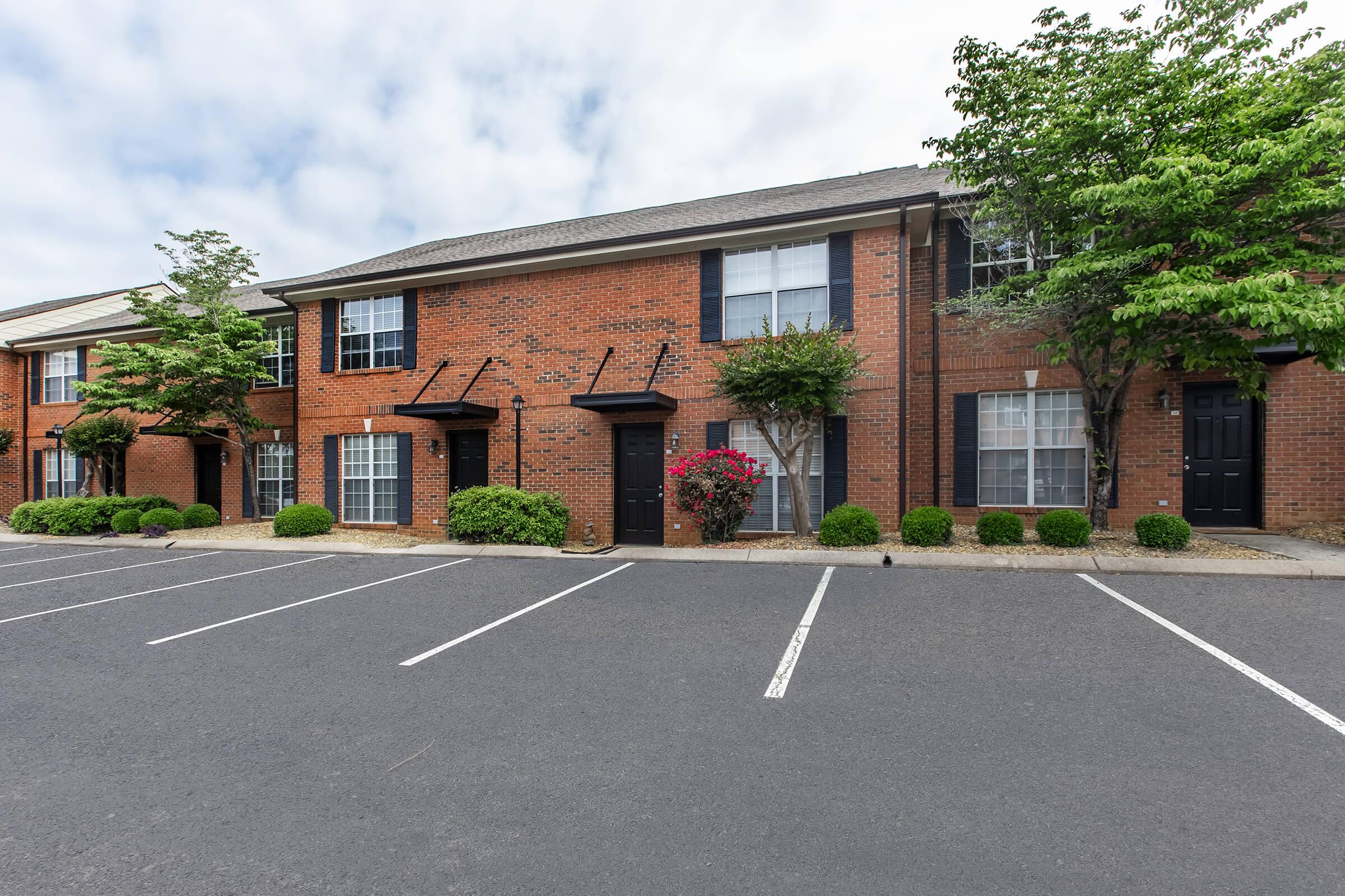 A row of brick apartment buildings with black doors and white windows. The landscaping features well-trimmed bushes and flowering plants, while a paved parking lot with empty spaces is in the foreground. The sky is partly cloudy.