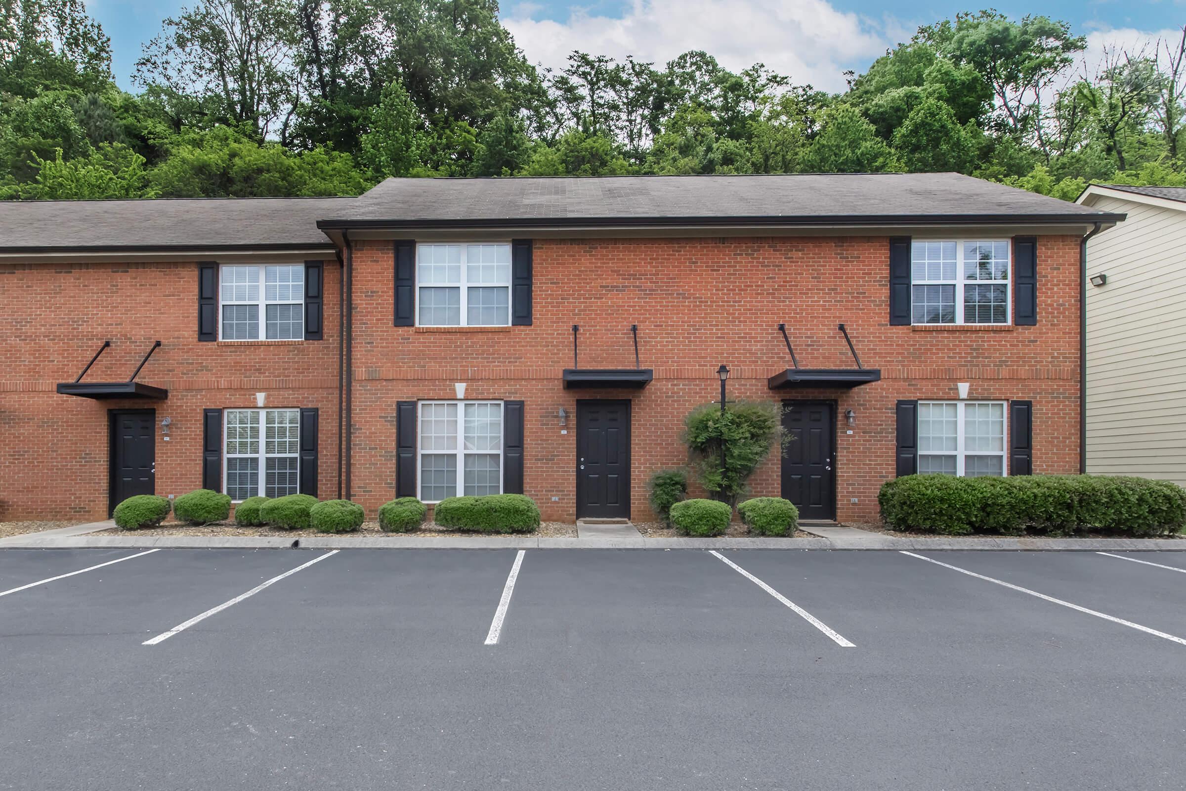 Two-story brick apartment building featuring four units with black shutters, bordered by neatly trimmed bushes. The parking area in front has clearly marked white stripes. Lush trees and greenery are visible in the background under a partly cloudy sky.