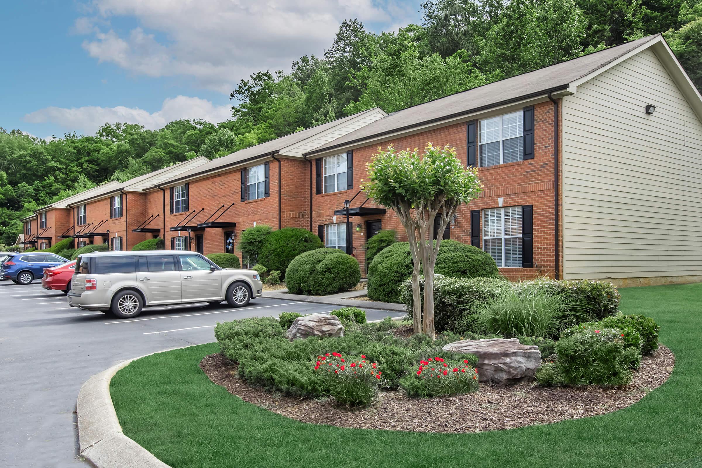 View of a residential apartment complex featuring brick buildings with landscaping, including shrubs and flower beds. In the foreground, a circular garden with red flowers and rocks is visible, alongside a well-maintained parking lot with several parked vehicles. The background showcases green trees and a blue sky.