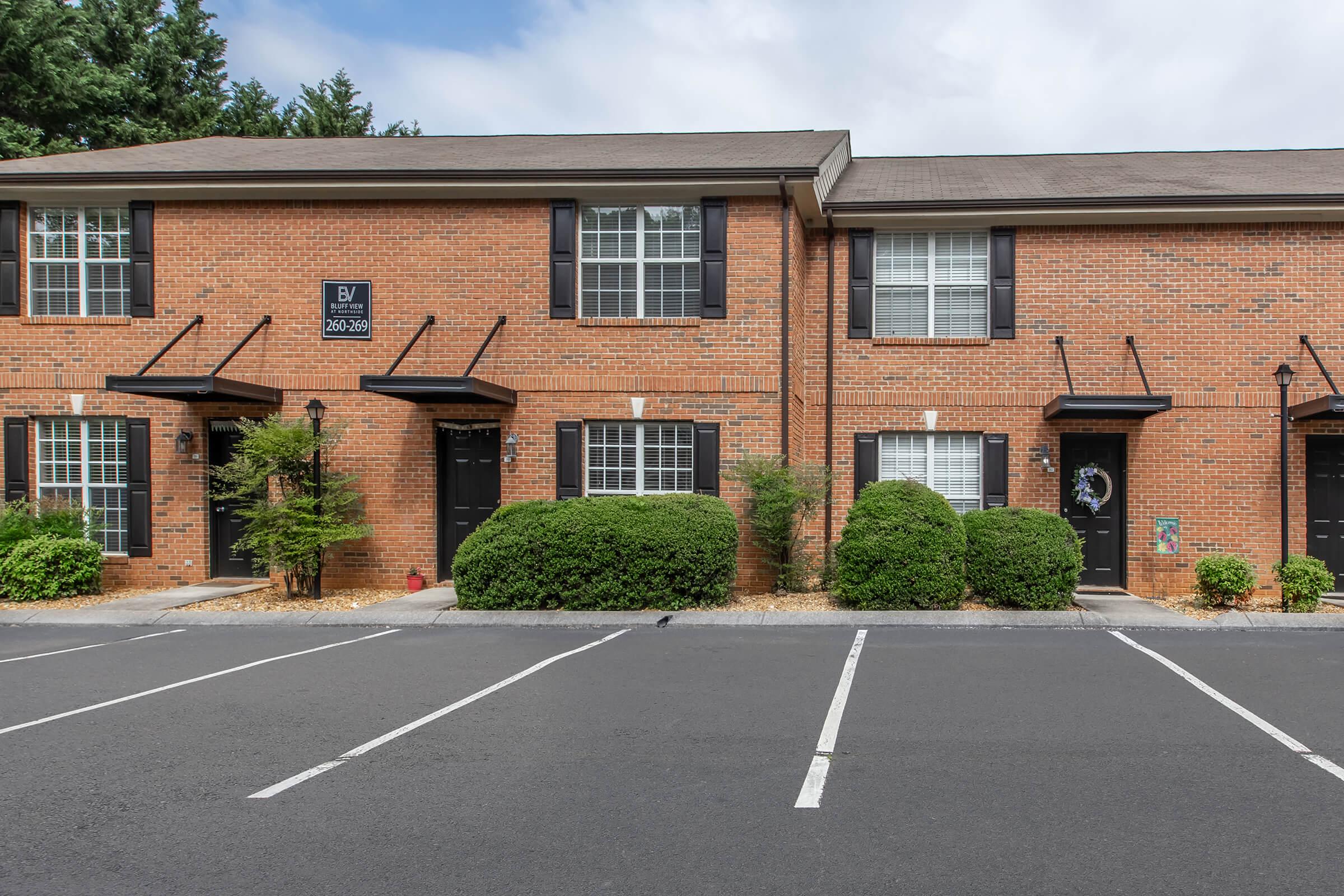 A view of a brick townhouse complex with multiple units. Each unit has black doors and white windows with black shutters. Neat shrubs are spaced along the front of the building. The parking area is visible in the foreground, with several empty parking spaces. The sky is partly cloudy.