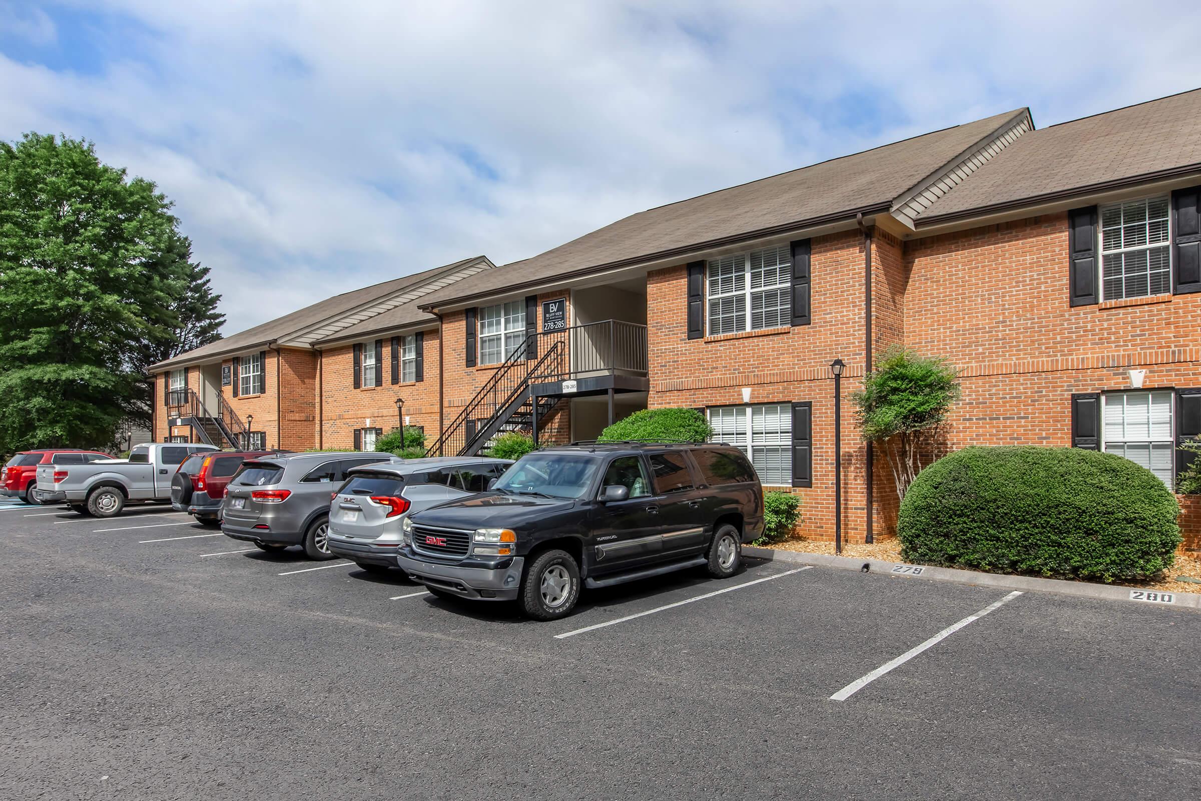 A row of brick apartment buildings with multiple parked cars in front. The buildings have stairs leading to upper units, and there are well-maintained shrubs and trees in the landscaping. The sky is partly cloudy, creating a pleasant atmosphere.