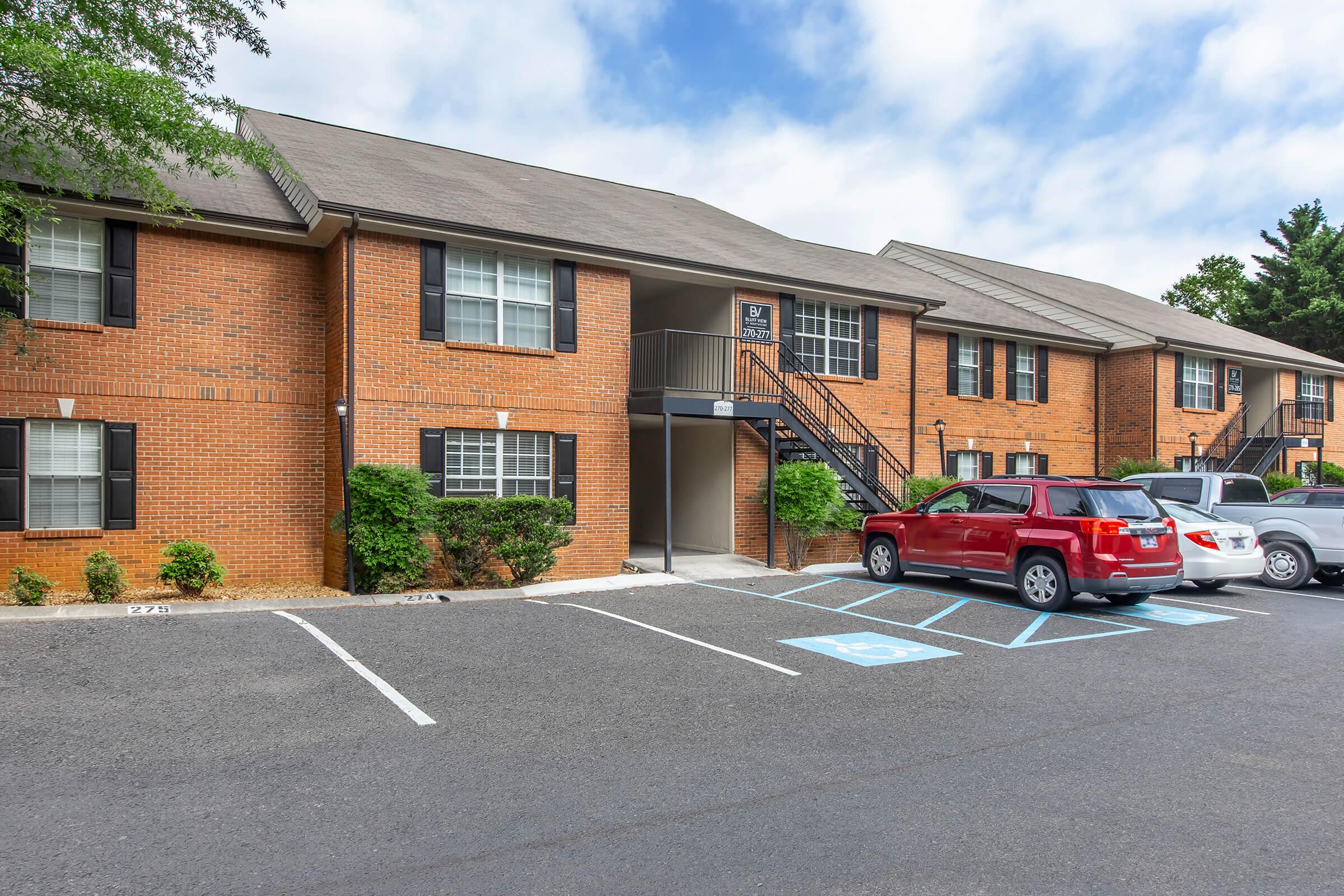 A brick apartment building featuring two levels with staircases leading to the upper units. The front has several parking spaces, including some marked for handicap accessibility. The exterior is well-maintained with shrubs and a clear sky in the background.