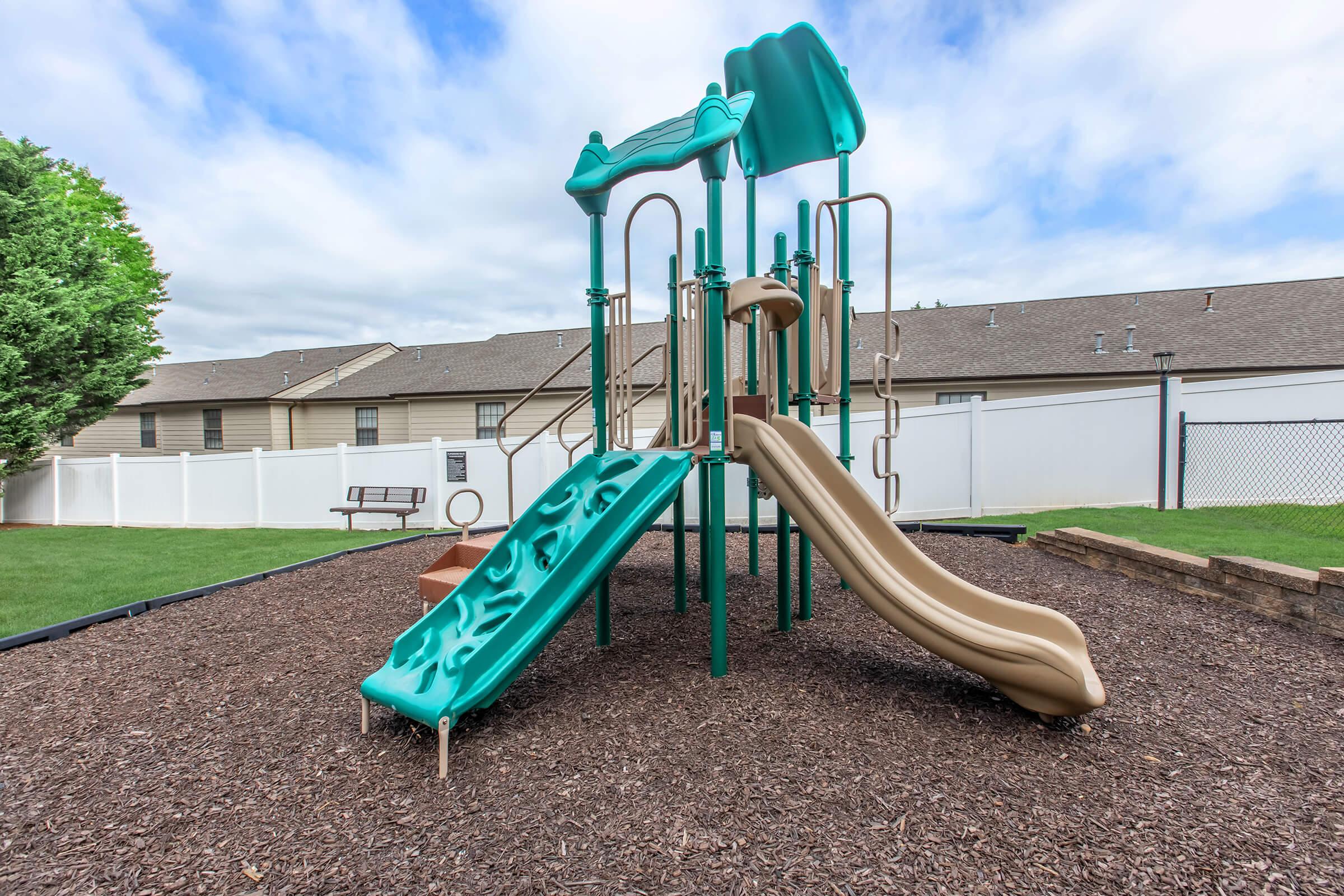 A playground structure featuring a green and tan slide, climbing walls, and multiple levels, set on a surface of wood chips. In the background, there are residential buildings and a white fence, along with a bench on grass. The sky is partially cloudy.