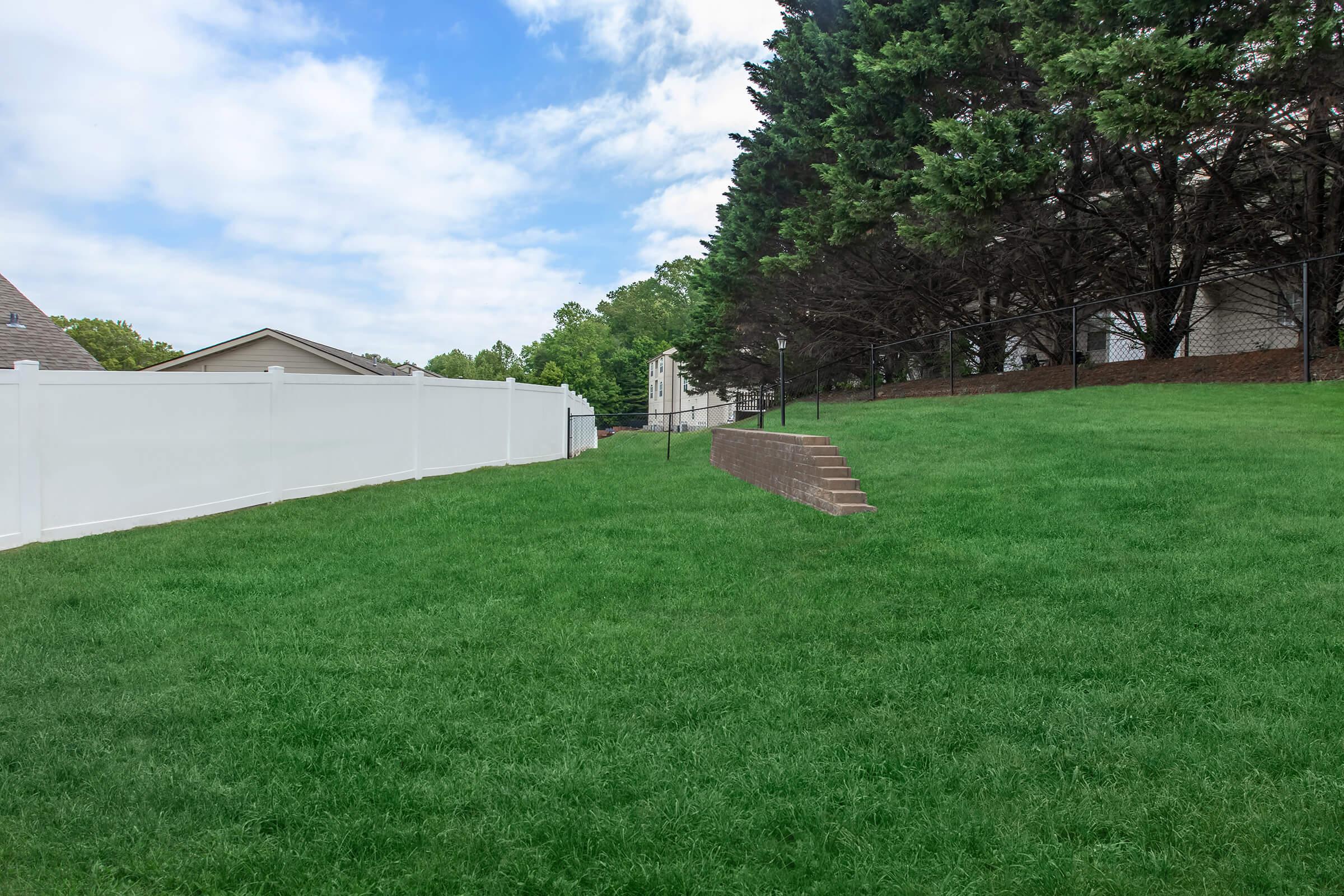 A grassy yard with a white fence on one side and a row of trees in the background. Stairs lead up to a higher level of the yard, with houses visible in the distance under a partly cloudy sky. The scene is serene and well-maintained.