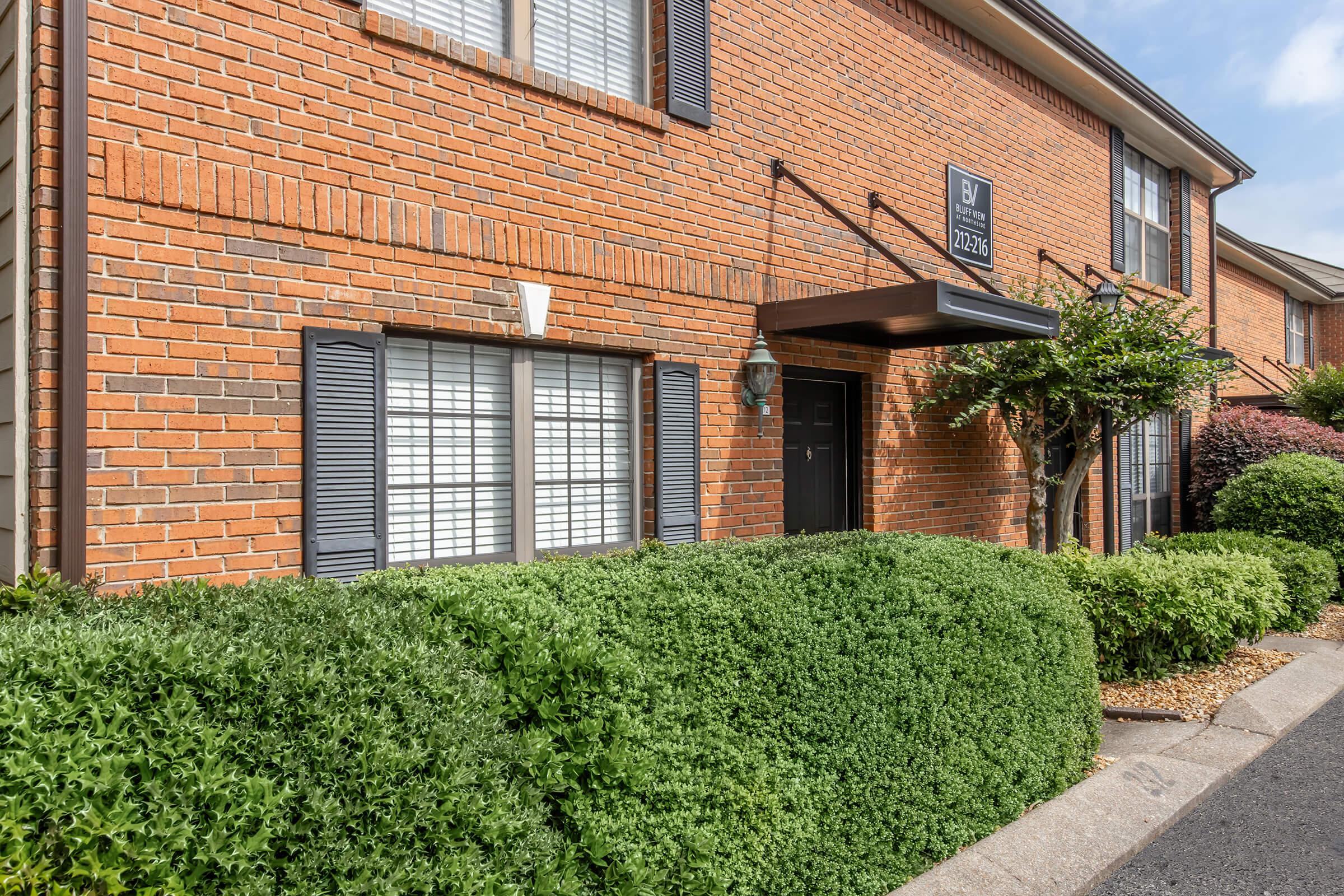 Exterior view of a brick building featuring a black entrance awning, large windows with shutters, and neatly trimmed green bushes in front. The scene is well-lit, showcasing the building's architectural details and landscaping.