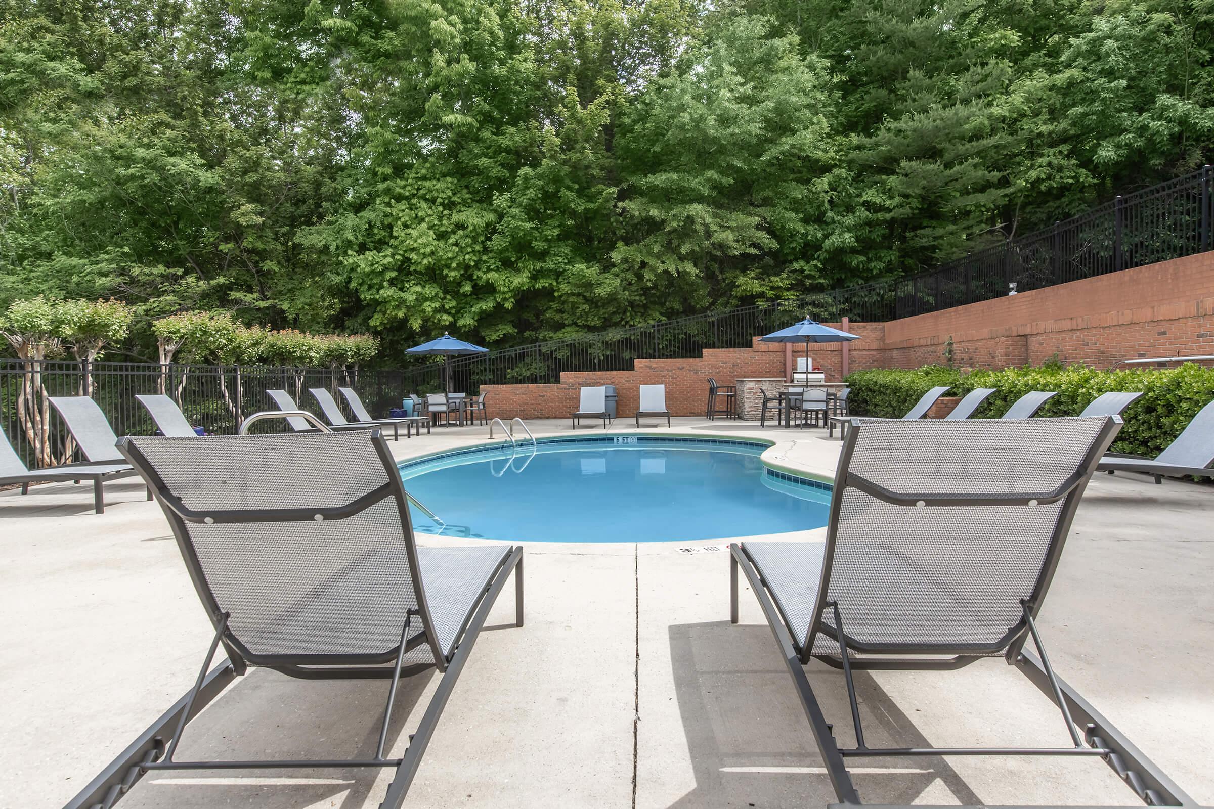 A serene pool area featuring a circular swimming pool surrounded by lounge chairs. The setting includes an enclosure of lush green trees and shrubs, with several blue umbrellas providing shade. The pool deck is clean and inviting, perfect for relaxation in a tranquil outdoor environment.