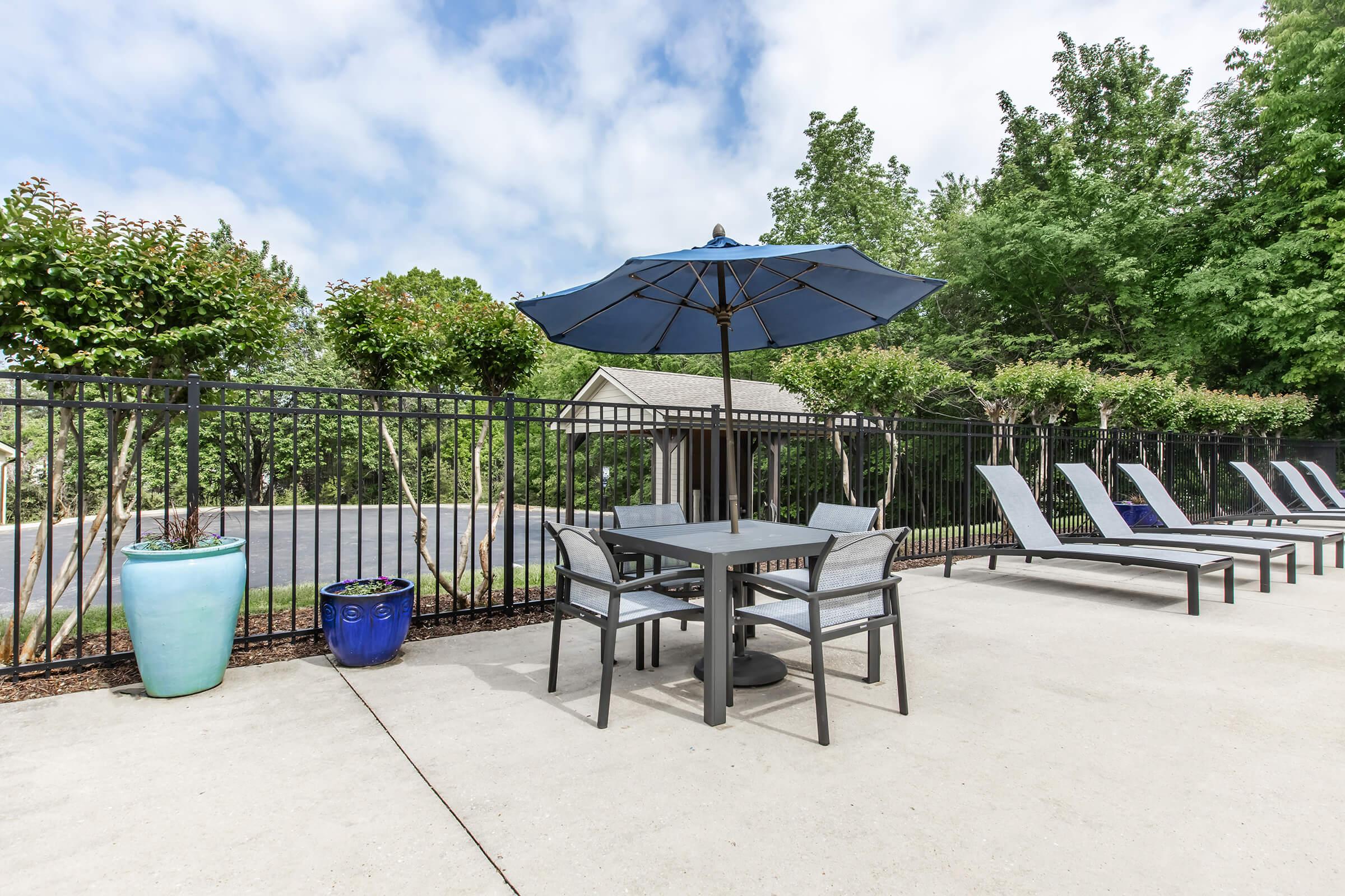 A serene outdoor space featuring a table and chairs under a blue umbrella, surrounded by lounge chairs. Lush greenery and decorative planters add to the tranquil atmosphere. The area is fenced and has a paved surface, ideal for relaxation.