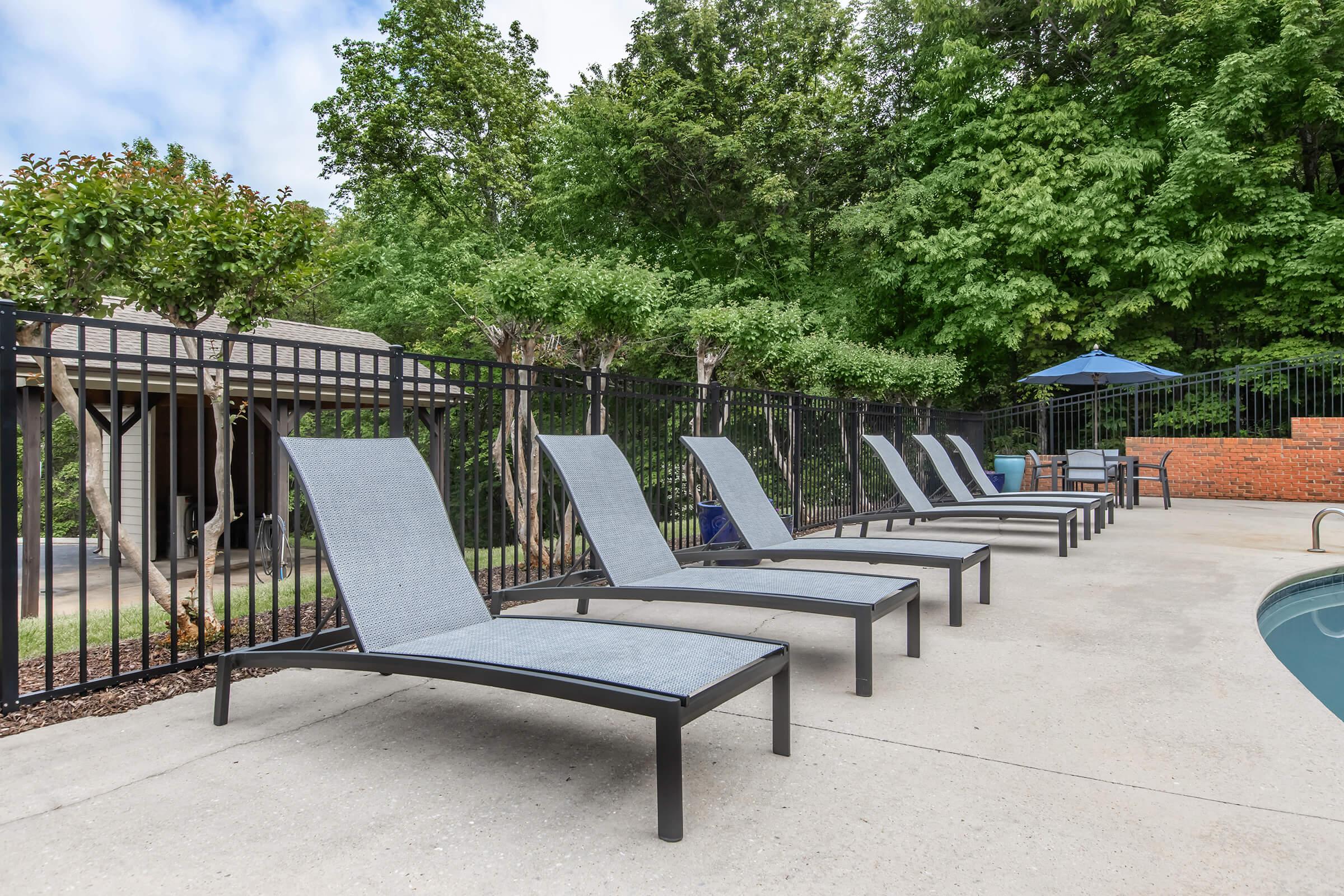 A tranquil outdoor space featuring multiple gray lounge chairs lined up beside a swimming pool. The area is surrounded by lush green trees and a black metal fence, with an umbrella providing shade nearby. The setting evokes a relaxing atmosphere ideal for leisure and sunbathing.