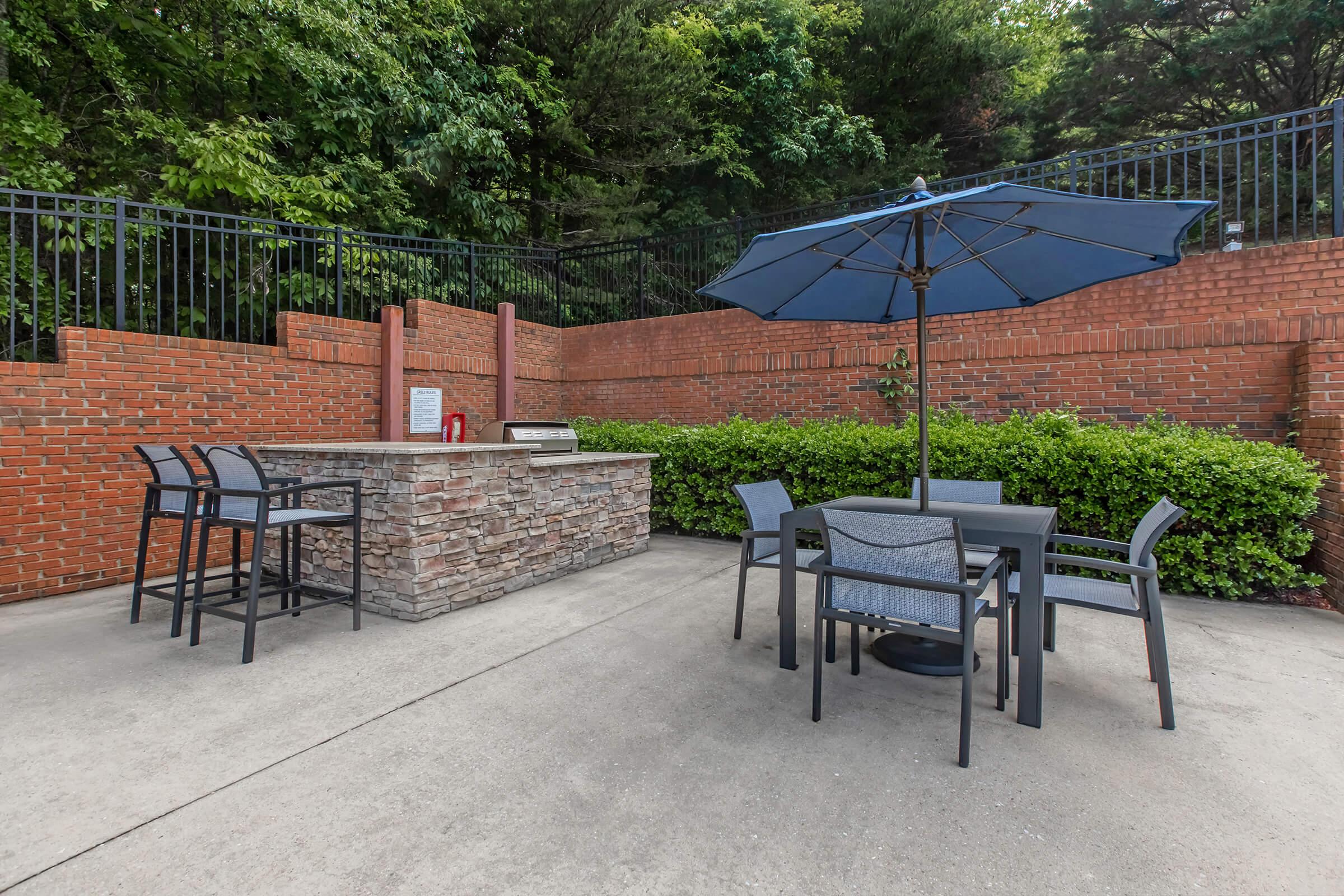 Outdoor patio area featuring a stone grill station and seating. There are several black dining chairs and tables arranged under a large blue umbrella. The space is bordered by green bushes and a brick wall, with trees in the background providing shade.