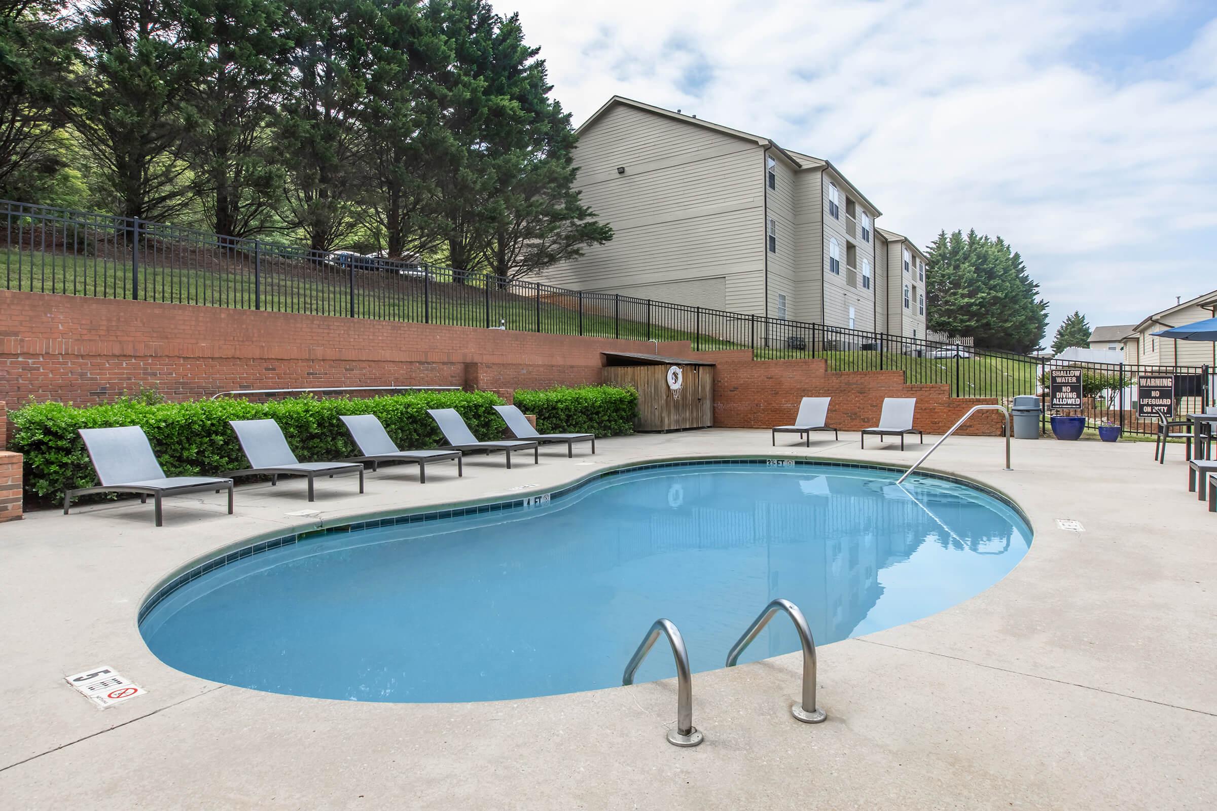 A serene outdoor swimming pool surrounded by lounge chairs, set against a backdrop of greenery and apartment buildings. The pool has a curved shape and a shallow entry, with a clear blue sky above. The area is well-maintained, offering a relaxing atmosphere for residents.