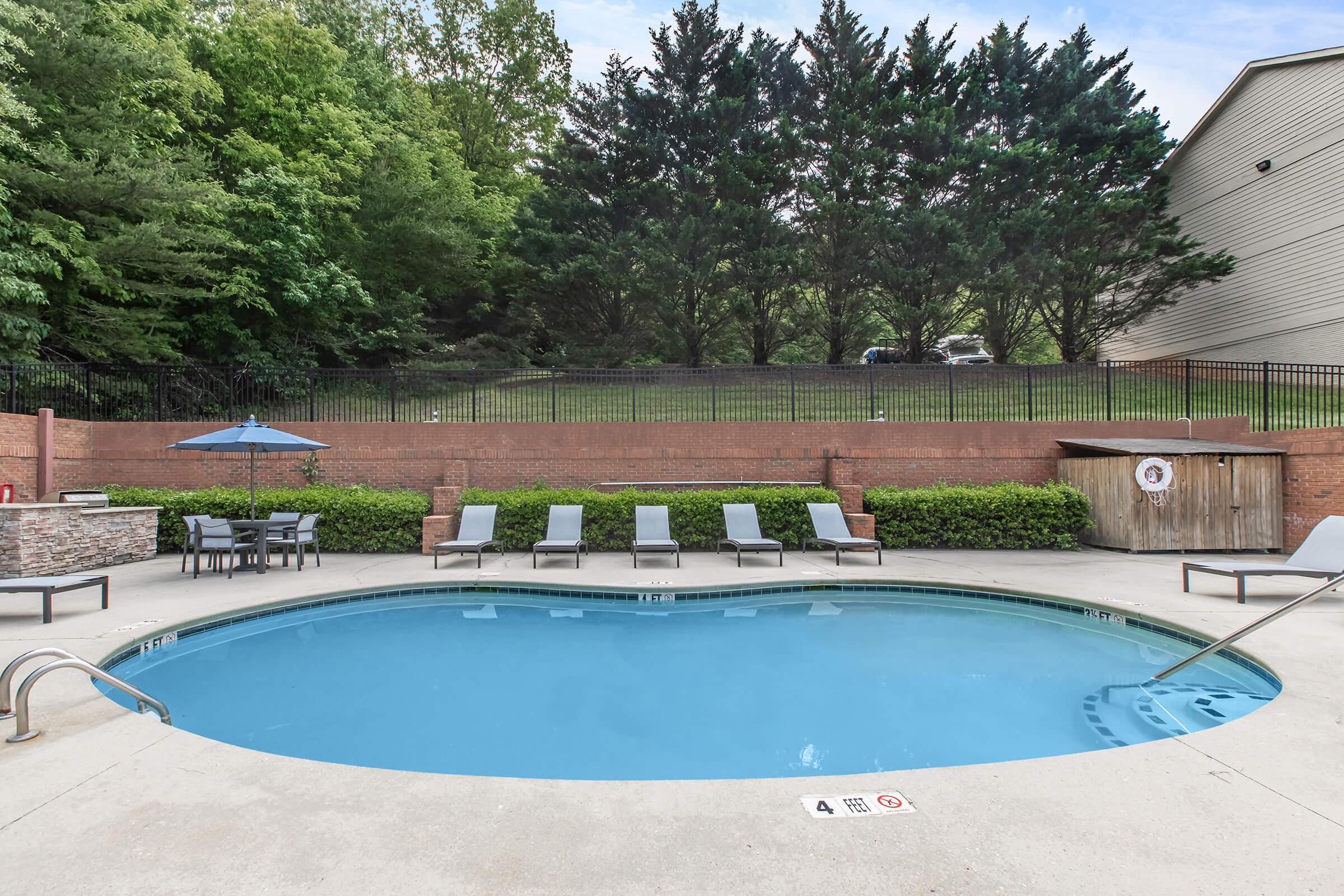 A swimming pool surrounded by lounge chairs, a patio area with tables, and an umbrella. Green trees and a grassy area are visible in the background, along with a wooden storage shed near the pool. The scene conveys a relaxing outdoor atmosphere.