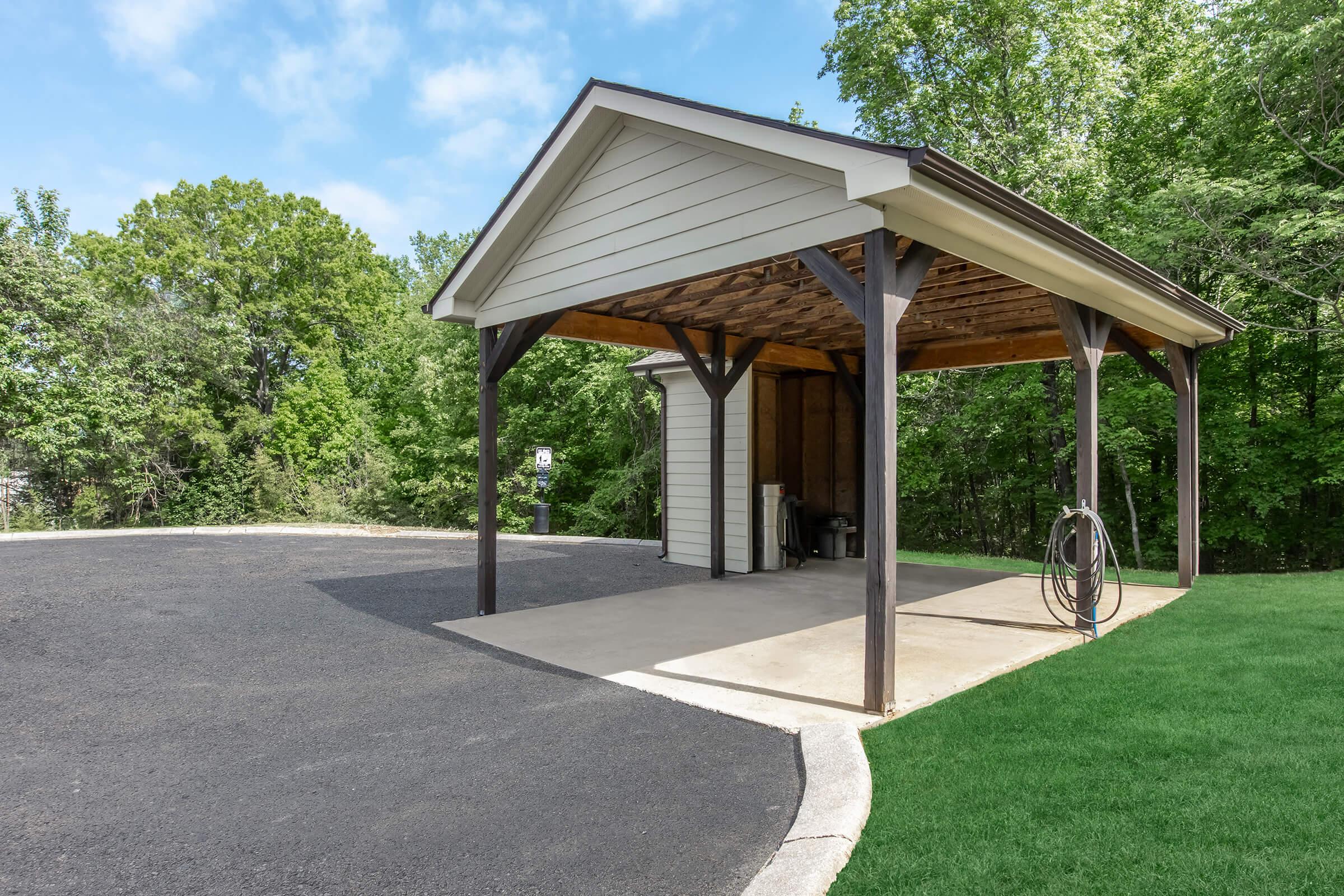 A covered carport with a concrete floor and wooden beams, surrounded by green trees. There is a paved driveway leading up to the carport, and a grassy area nearby. The sky is partly cloudy, creating a bright, inviting atmosphere.