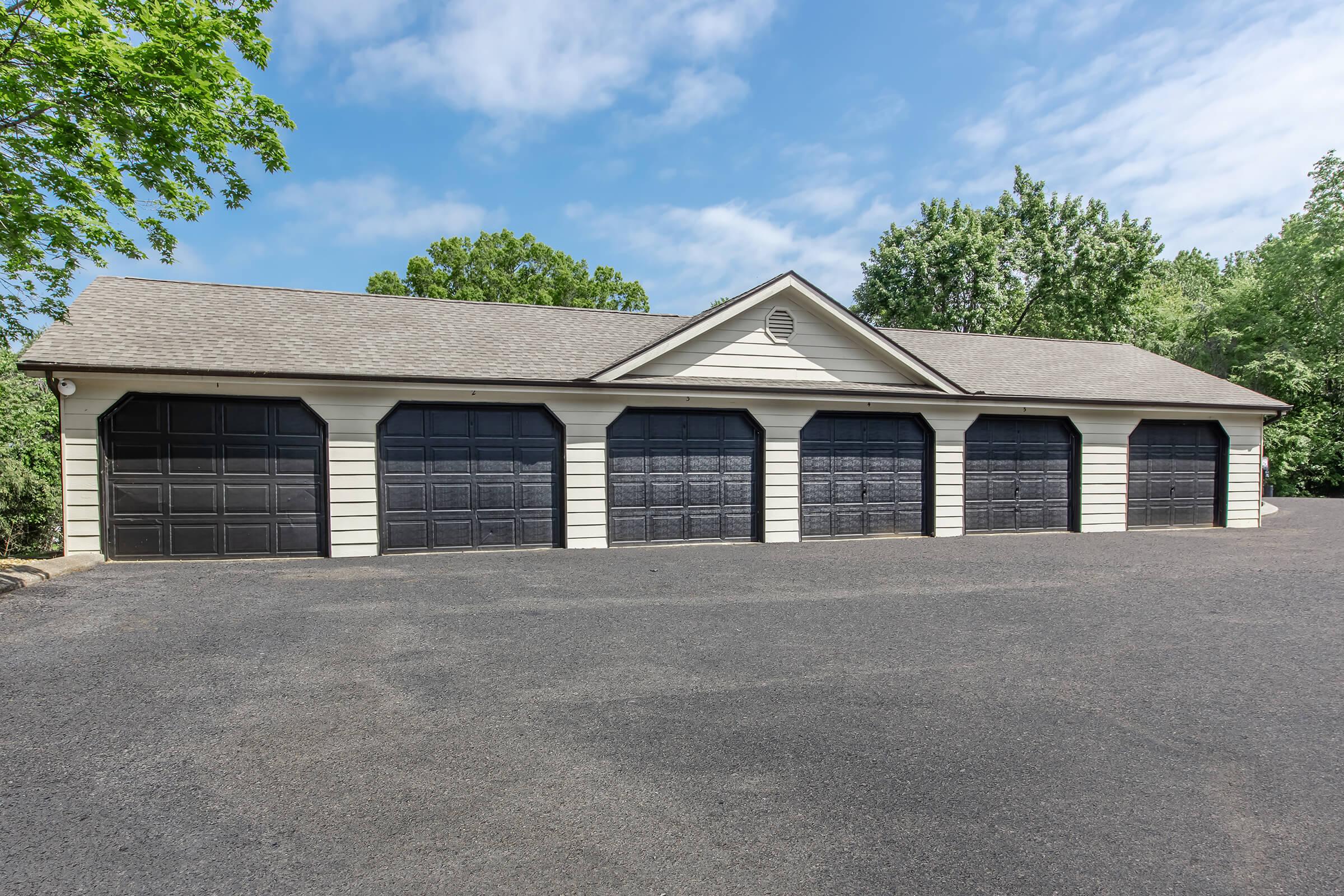 A row of five black garage doors on a light-colored building, situated on a paved driveway. The surrounding area features trees and a clear blue sky, creating a serene setting.
