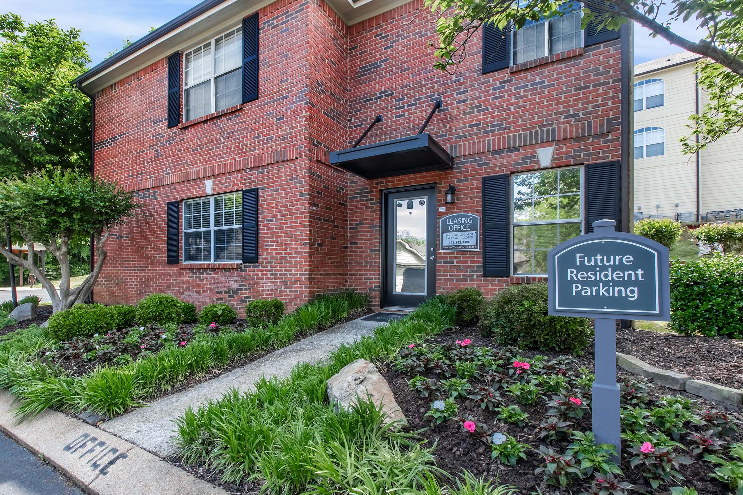 A red brick building labeled as the leasing office, featuring large windows and a welcoming entrance. The front yard has neatly trimmed shrubs, colorful flowers, and a sign indicating "Future Resident Parking." The overall setting is well-maintained and inviting.
