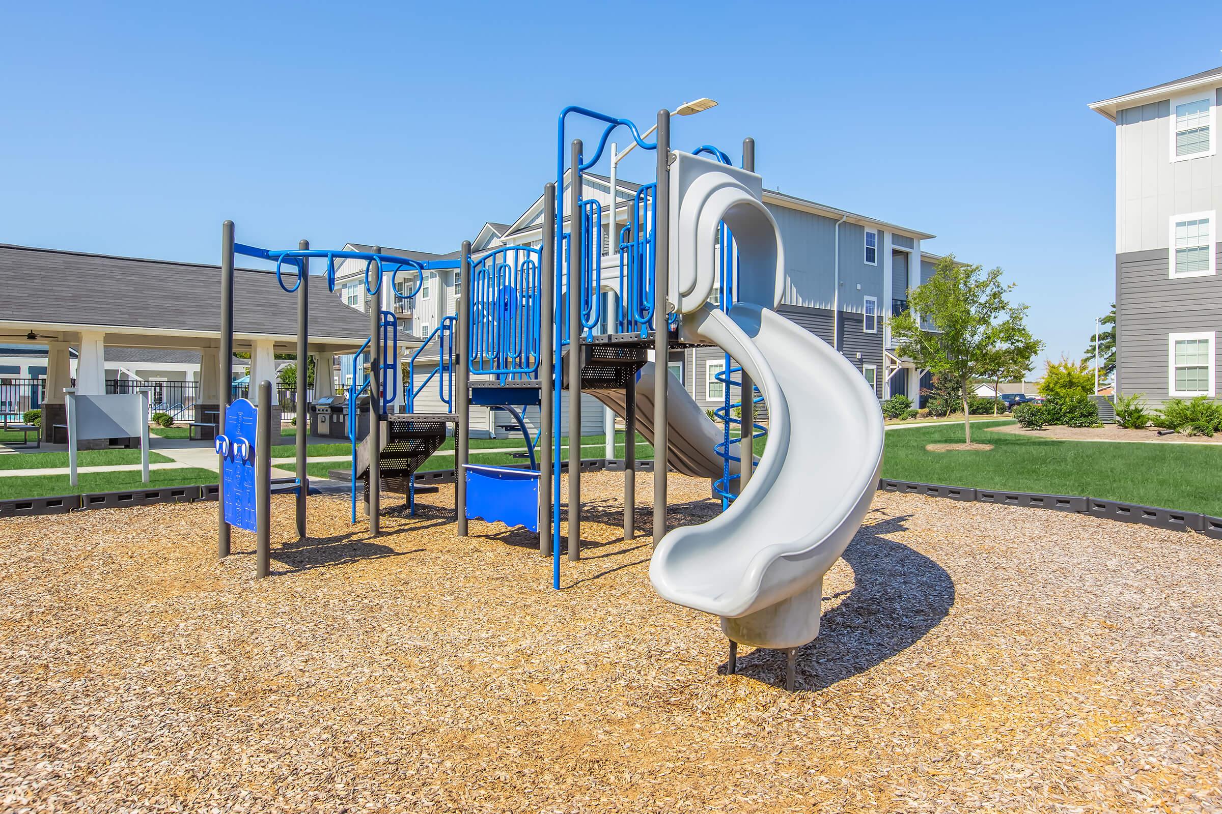 Children's playground featuring a blue and silver play structure with a slide. The ground is covered in wood chips, and nearby are buildings with a grassy area. The sky is clear and sunny, creating a vibrant outdoor activity space.