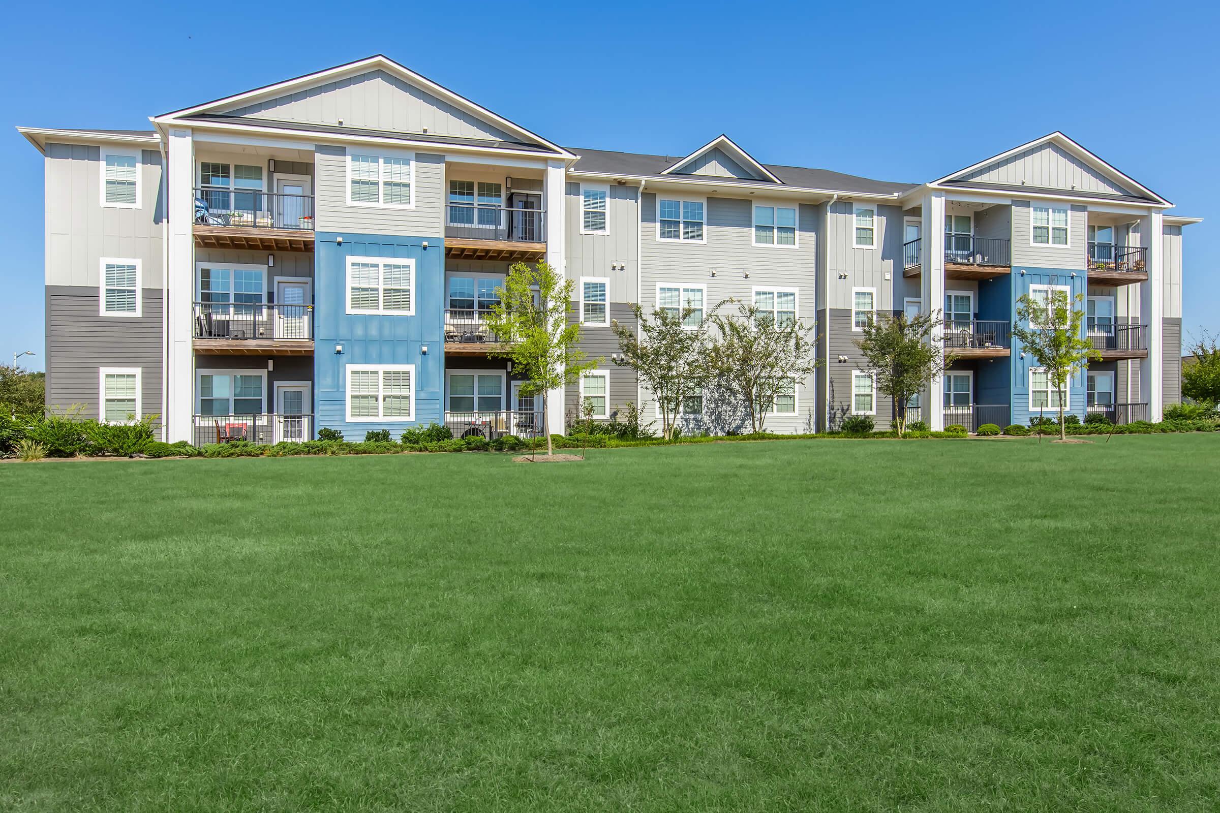 Modern multi-story apartment building with a mix of gray and blue siding, featuring balconies and large windows. The building is surrounded by well-maintained green grass and small trees under a clear blue sky.