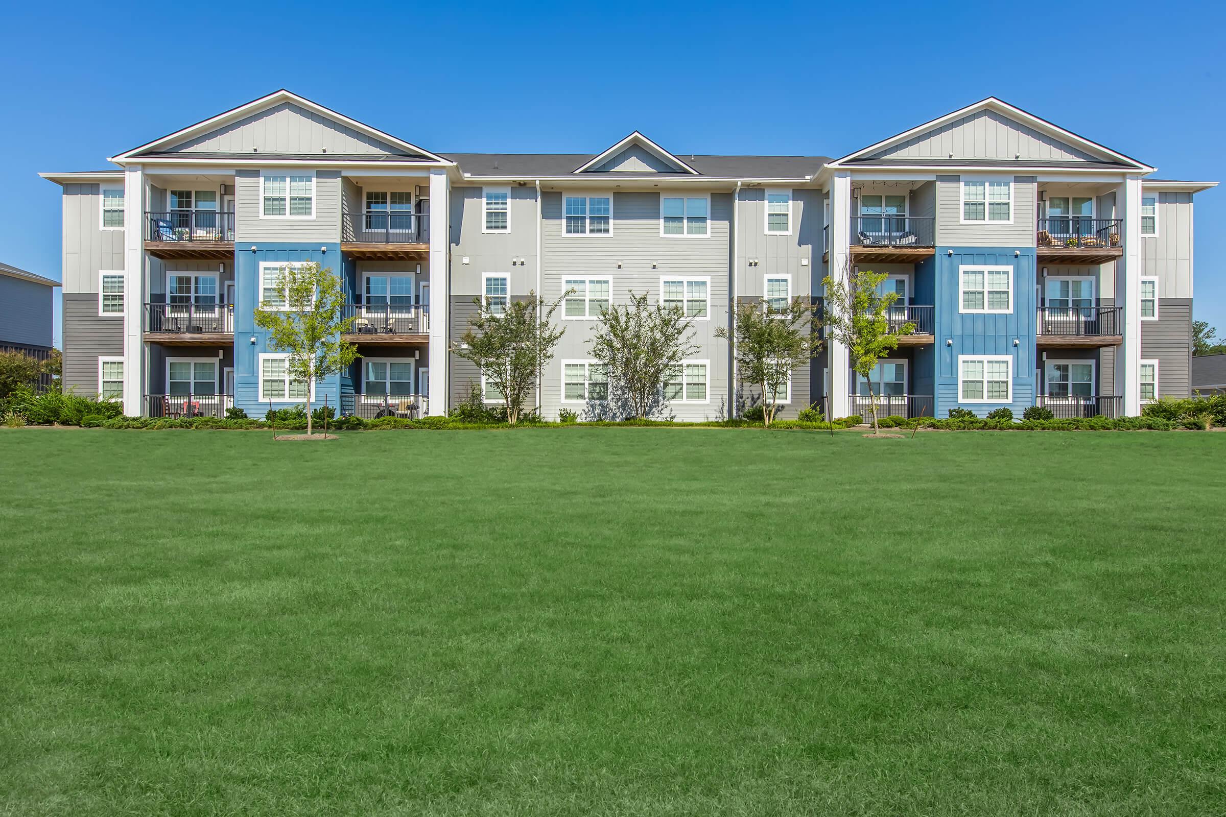A three-story residential apartment building with a mix of light blue and gray exteriors. The building features multiple balconies and is surrounded by green grass and small trees under a clear blue sky.
