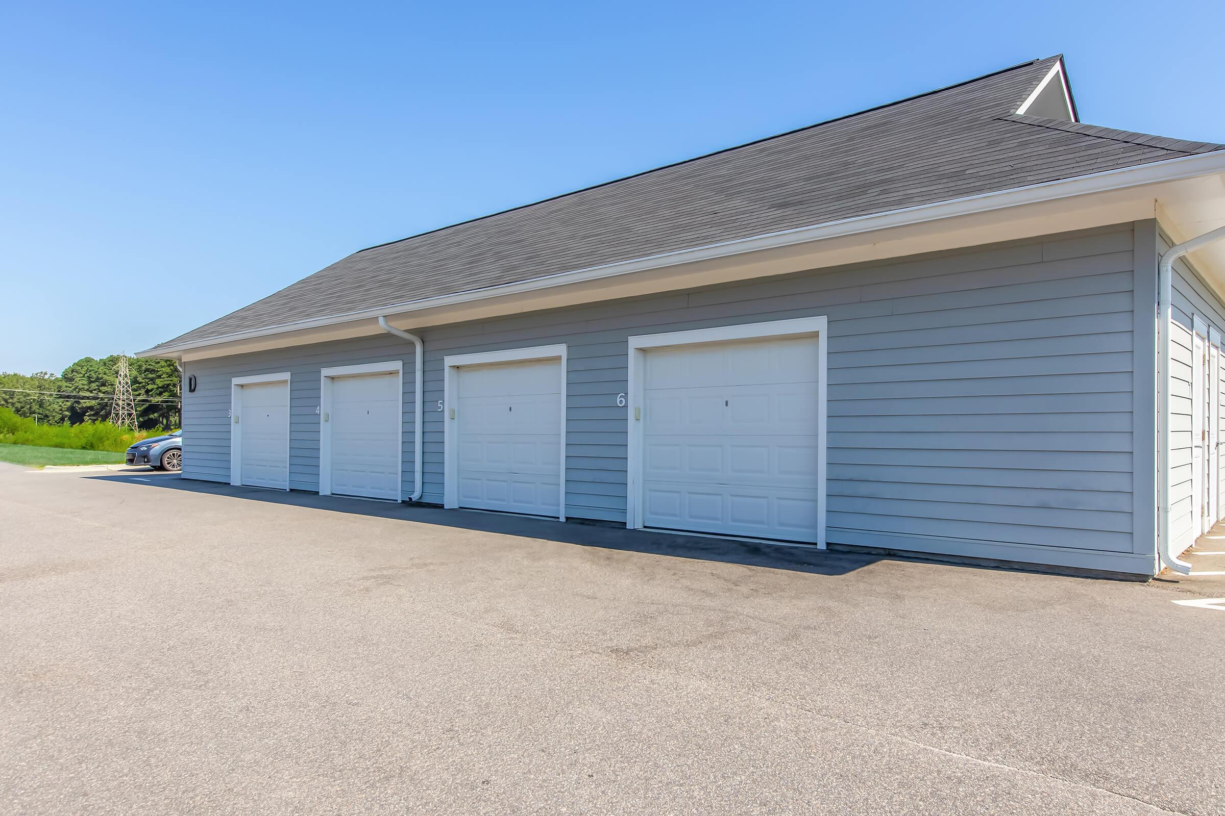 A row of gray storage garages with white doors under a clear blue sky. The pavement is well-maintained, and there's a green area in the background, suggesting a suburban or rural setting. The garages are numbered, with at least one door appearing half-open.
