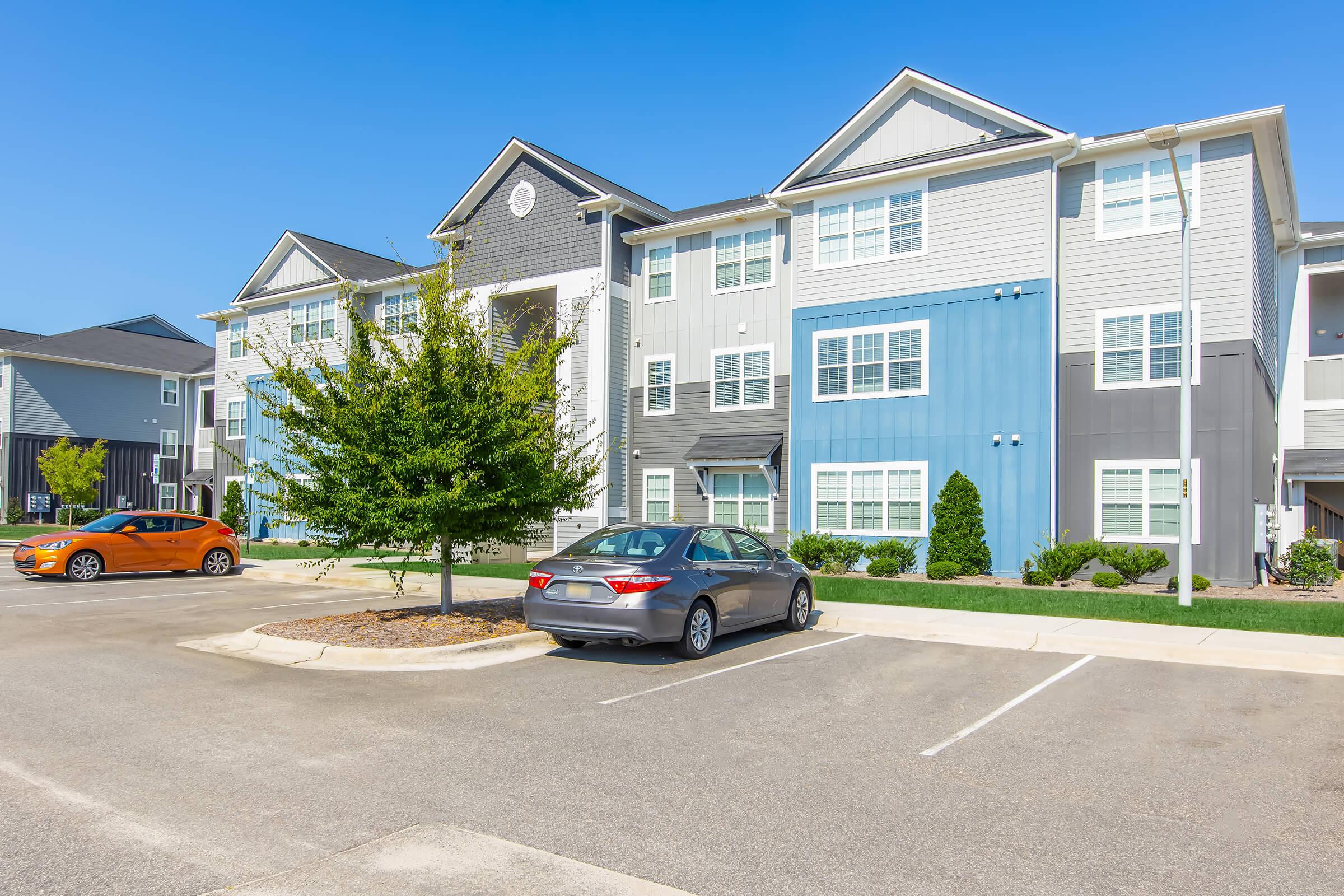Apartment complex with two buildings featuring a mix of grey and blue exteriors. A grey sedan is parked in front of a landscaped area with small trees. An orange car is parked nearby. The sky is clear and blue, indicating a sunny day.