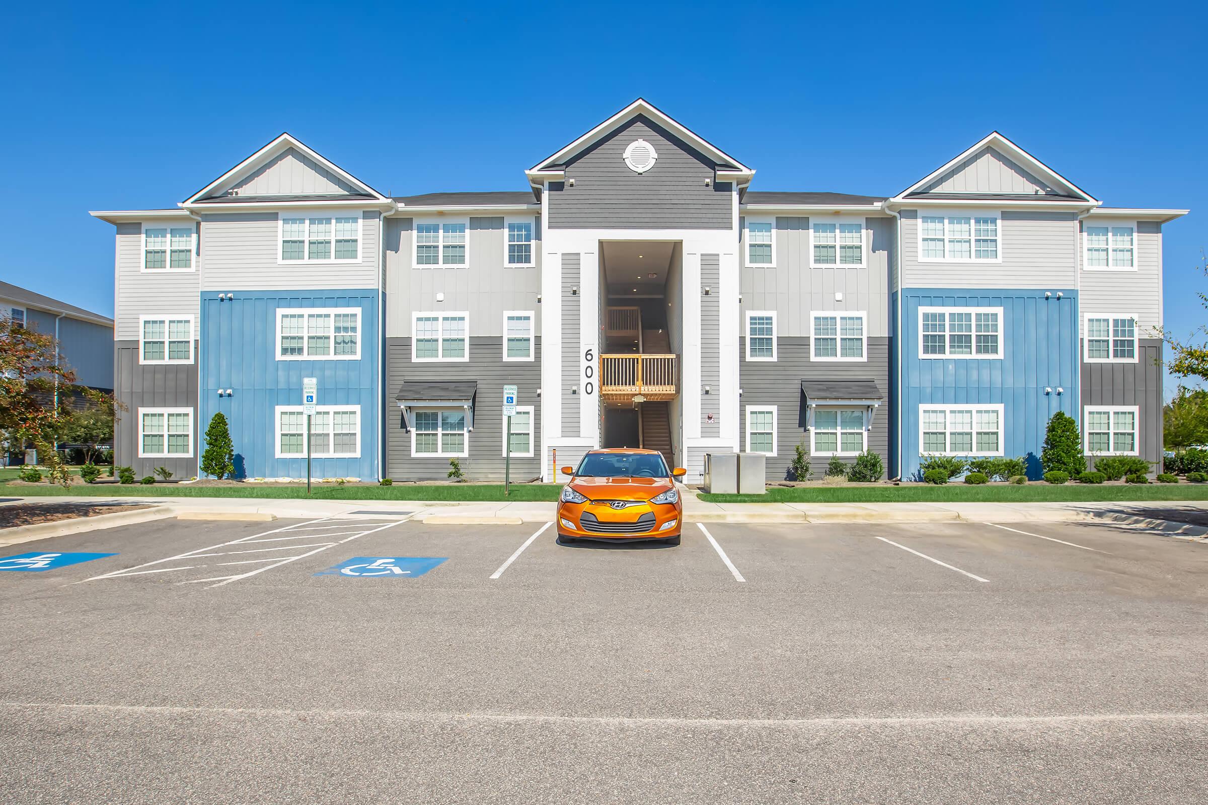 A modern, multi-story apartment building with a mix of blue and gray exterior. In the foreground, there's a bright orange car parked in an empty lot with several parking spaces visible, including designated handicap spots. The sky is clear and blue, enhancing the vibrant appearance of the building.