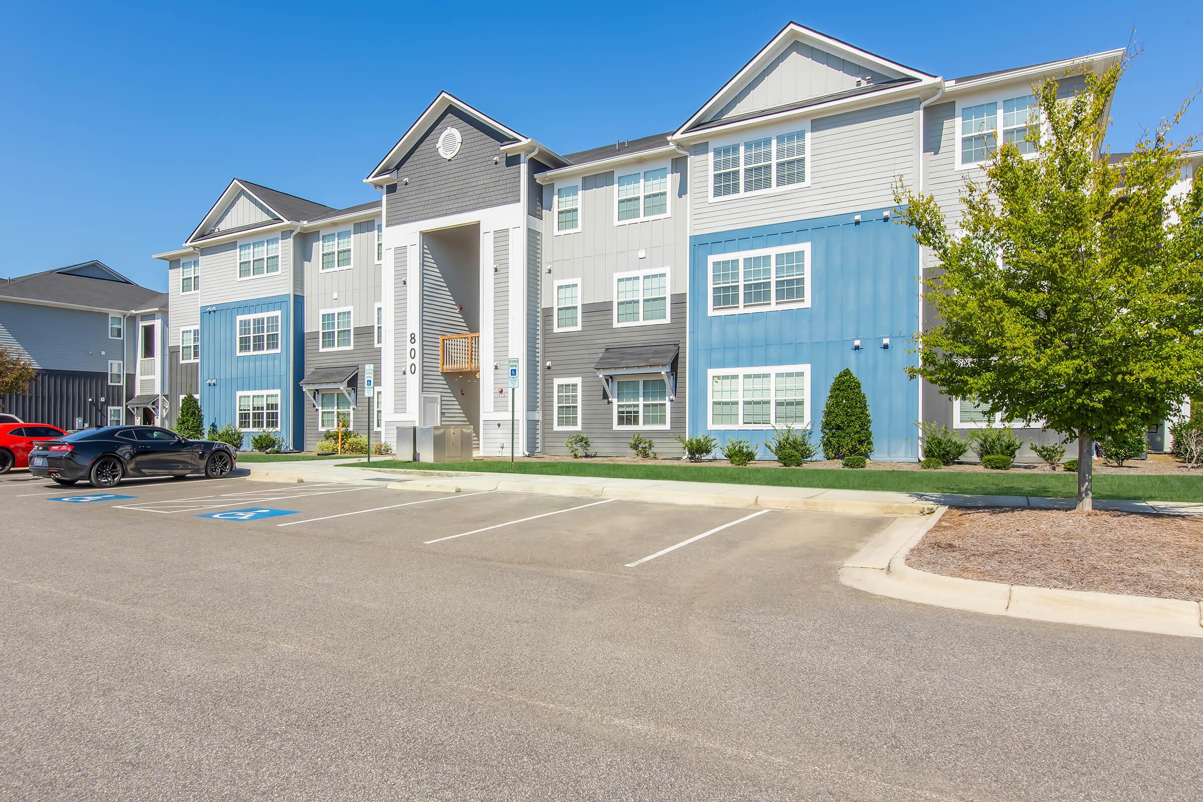 A modern apartment building featuring a mix of gray and blue exterior with multiple windows. In the foreground, there is a parking lot with marked spaces and a black sports car. Trees and landscaping are visible alongside the building, creating a welcoming environment. Bright, clear sky overhead.