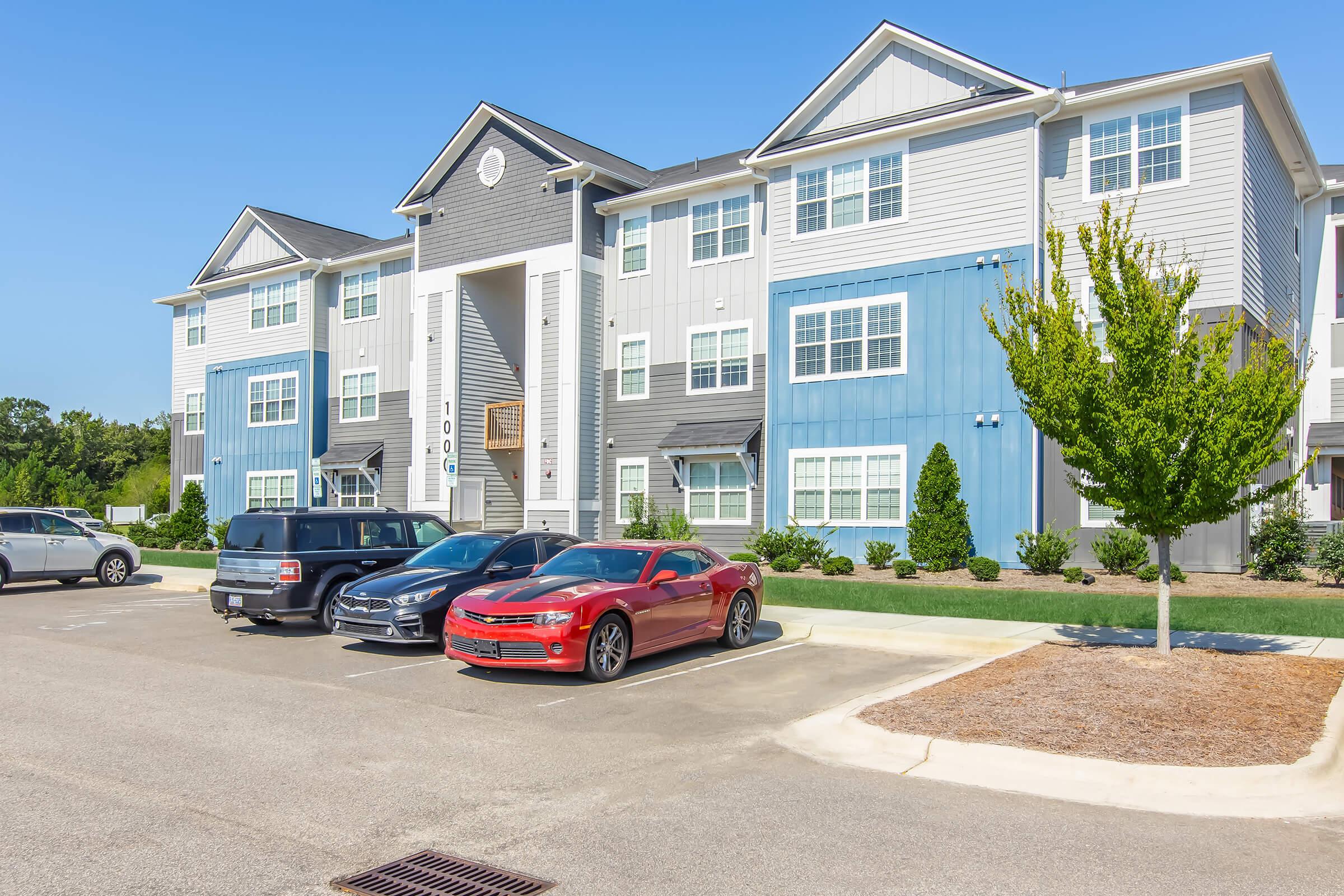 A modern multi-story apartment building with a mix of blue and gray siding. Several vehicles are parked in front, including a black SUV and a red sports car. The setting features landscaped areas with small trees and shrubs, and the sky is clear and sunny.