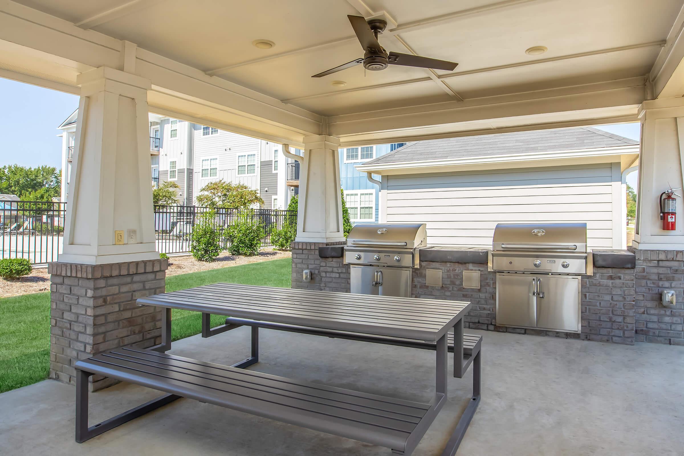 Covered outdoor grilling area with two stainless steel grills and a large gray picnic table. Located in a landscaped environment with green grass and nearby residential buildings. The space is well-lit with a ceiling fan for ventilation. Ideal for gatherings and barbecues.