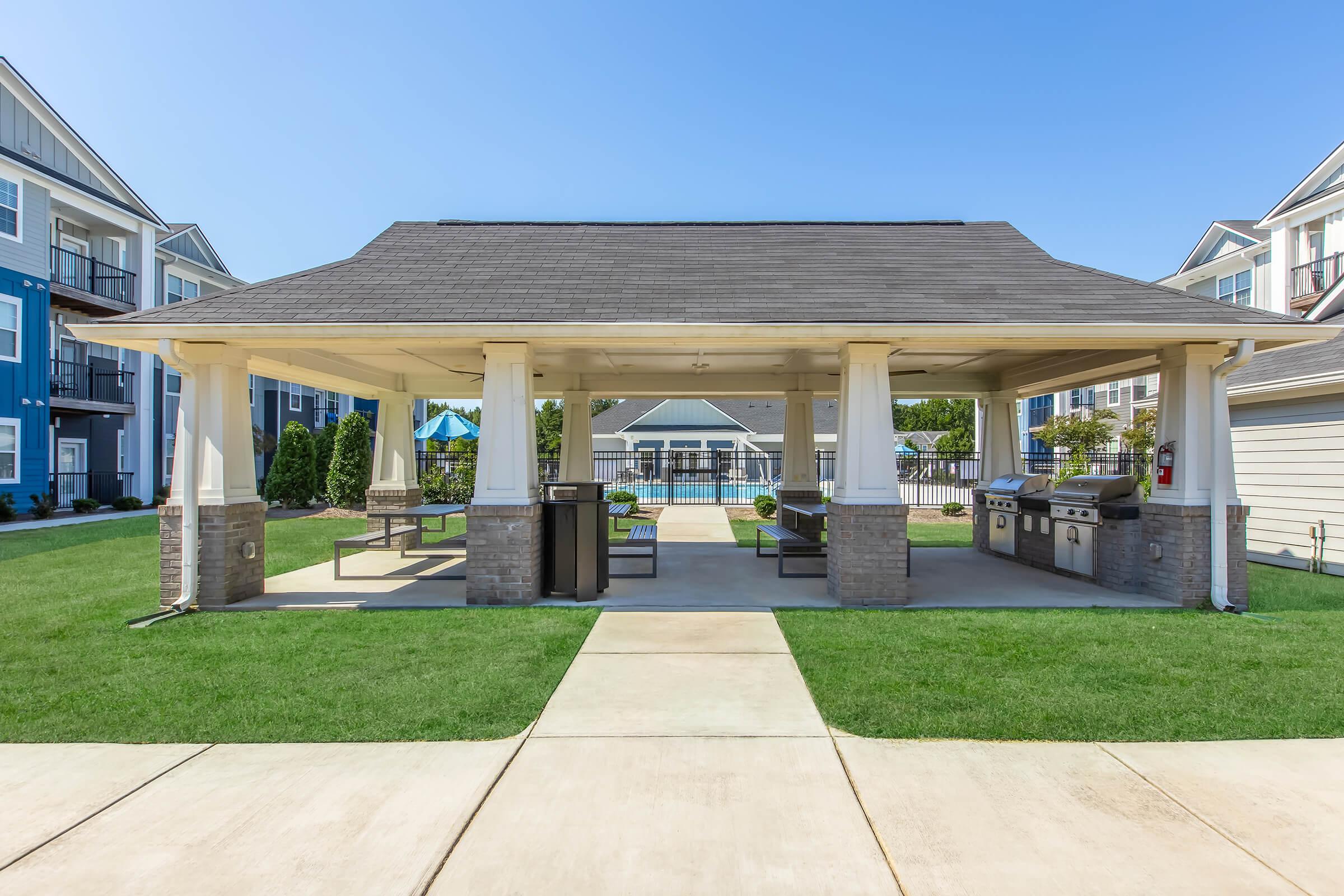 A spacious outdoor pavilion with a gabled roof and open structure, featuring multiple picnic tables and barbecue grills. Surrounding green grass and nearby residential buildings create a welcoming atmosphere on a sunny day.