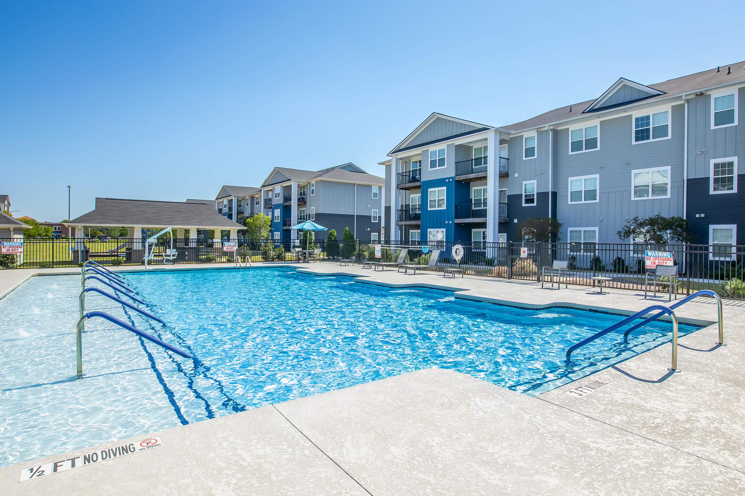 A clear blue swimming pool surrounded by a well-maintained area, featuring a shallow end with a "No Diving" sign. In the background, there are modern apartment buildings with balconies and a shaded seating area with umbrellas. The scene is bathed in bright sunlight against a clear blue sky.