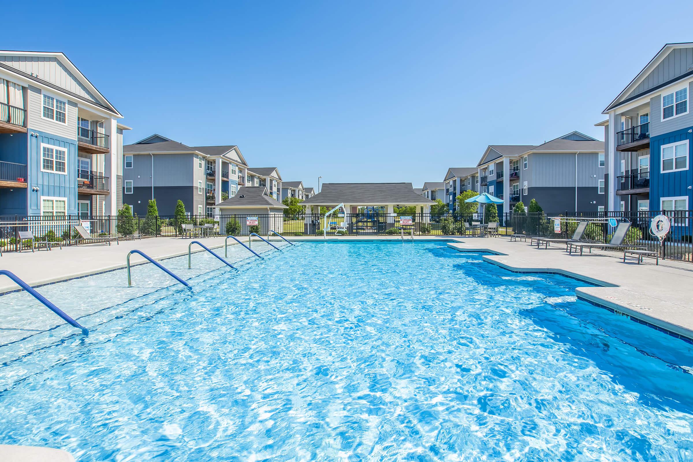 A large swimming pool with clear blue water, surrounded by modern apartment buildings. The pool area features lounge chairs and umbrellas, and a sunny blue sky is overhead. Green landscaping adds a fresh touch to the scene.