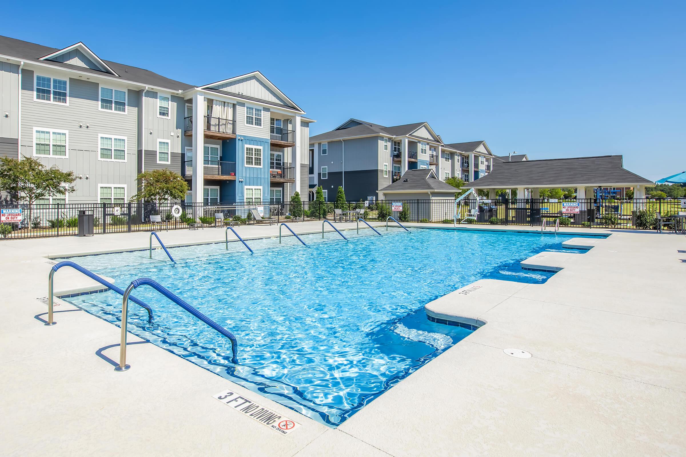A clear blue swimming pool surrounded by lounge areas and chairs, with an apartment complex in the background. The pool features steps leading in and is enclosed by a black railing and fencing. Bright, sunny weather with a clear blue sky enhances the inviting atmosphere.