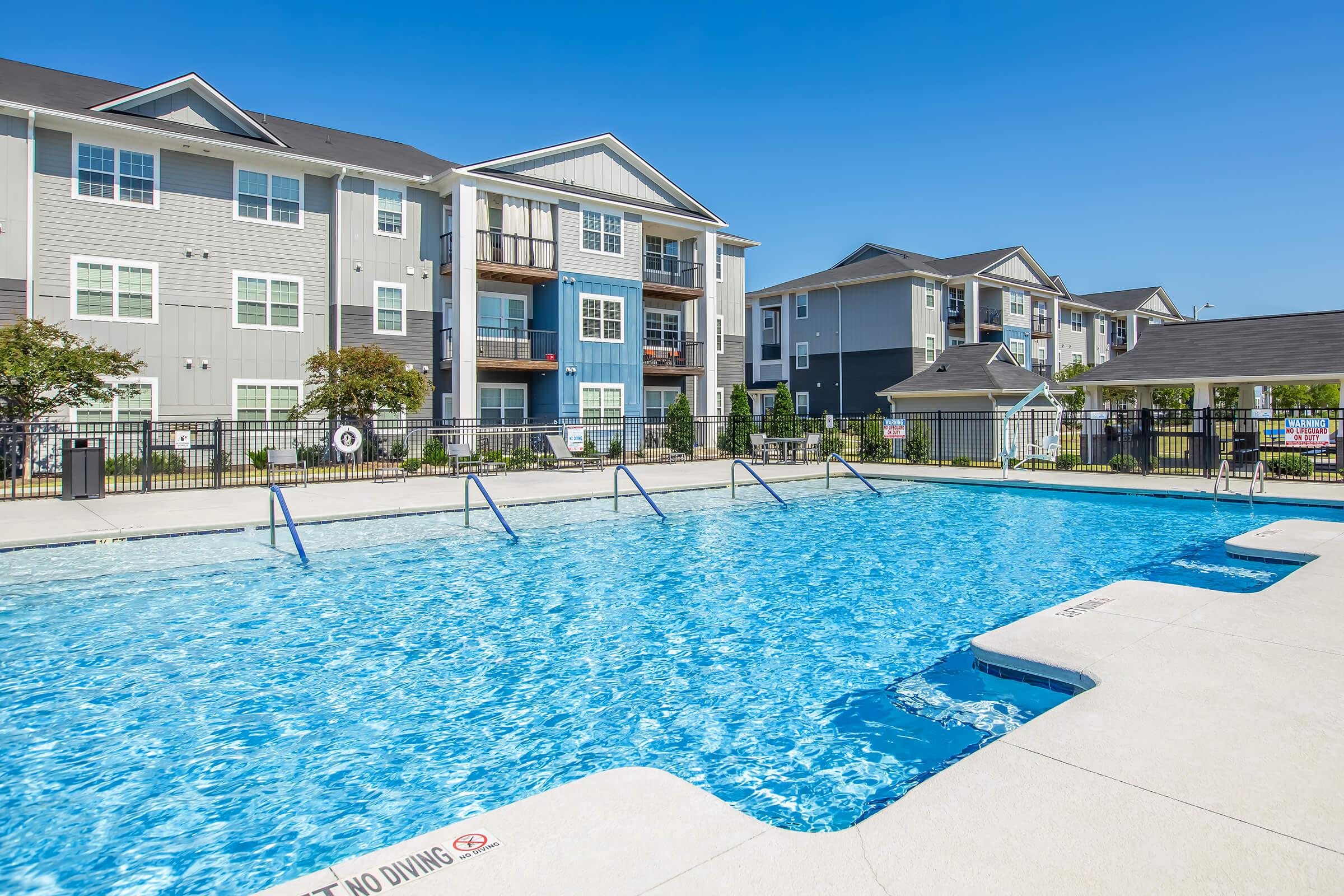 A clear blue swimming pool surrounded by a concrete deck, with fenced areas in the background featuring multi-story apartment buildings. The sky is bright and sunny, and there are signs indicating no diving. Lush greenery can be seen along the pool area.