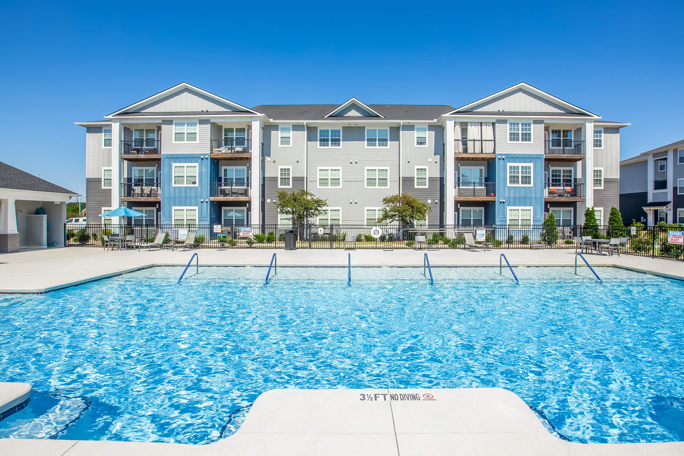 A bright and inviting apartment complex featuring a swimming pool in the foreground. The pool has multiple lanes and clear blue water, surrounded by lounge areas. The buildings in the background are modern, with balconies and a mix of gray and blue siding, under a clear blue sky.