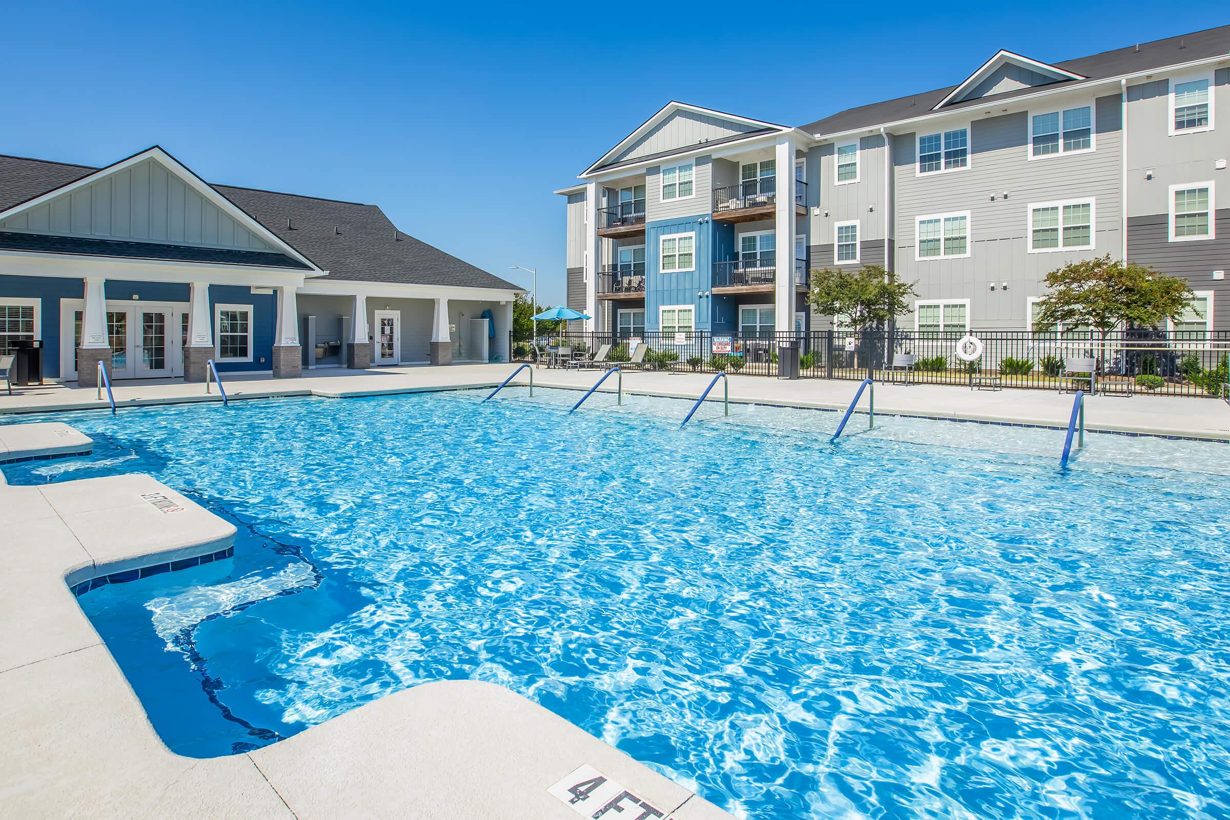 A clear blue swimming pool with diving lanes, surrounded by a paved deck. In the background, there are modern apartment buildings with a mix of gray and blue exteriors. The sky is bright and sunny, creating a cheerful atmosphere. Lush greenery is present around the pool area, adding to the inviting setting.