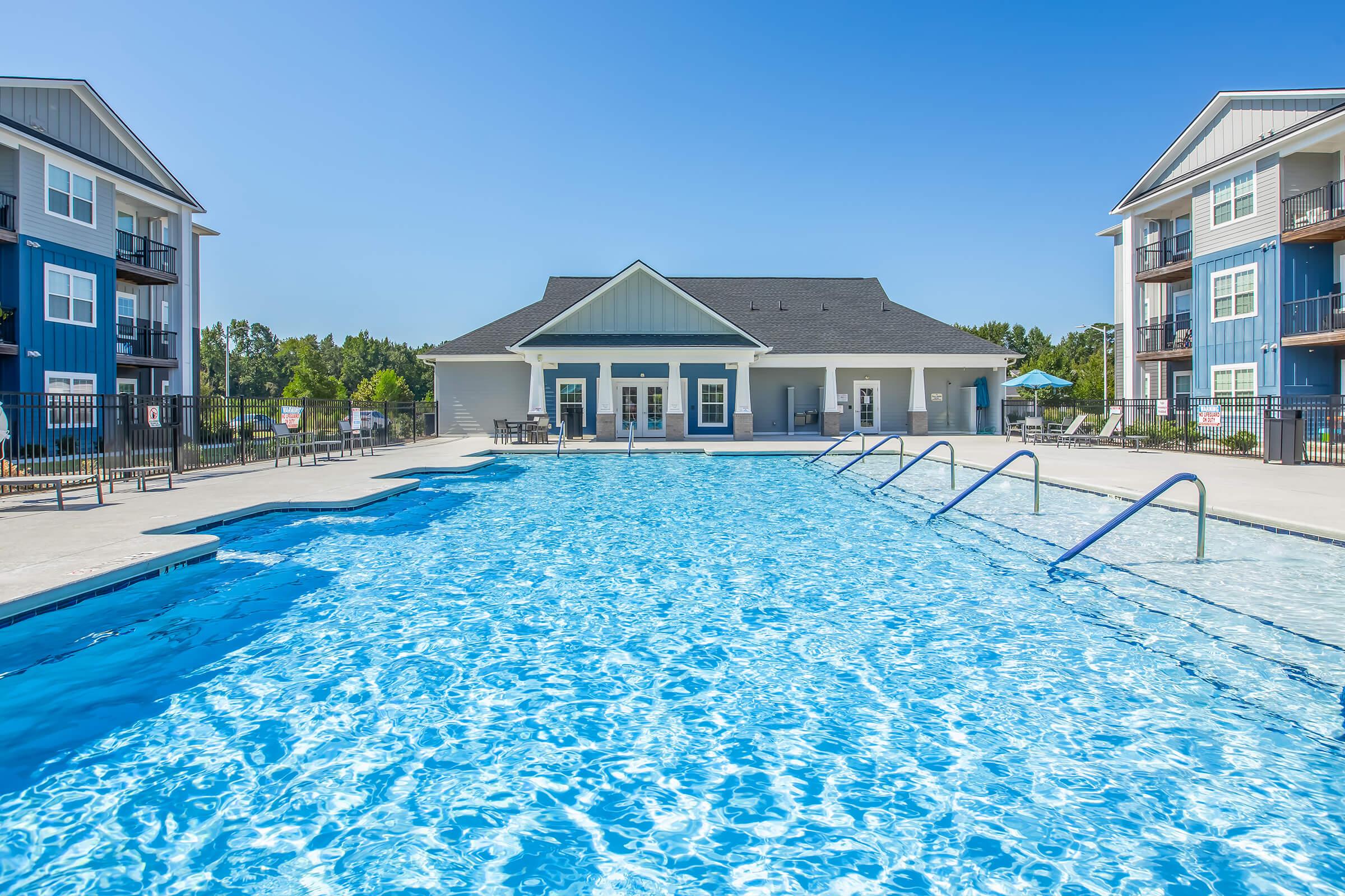 A clear blue swimming pool surrounded by modern apartment buildings under a bright blue sky. The pool features a textured surface and stairs leading into the water, with lounge chairs and a clubhouse in the background. Sunlight reflects off the water, creating a vibrant atmosphere.