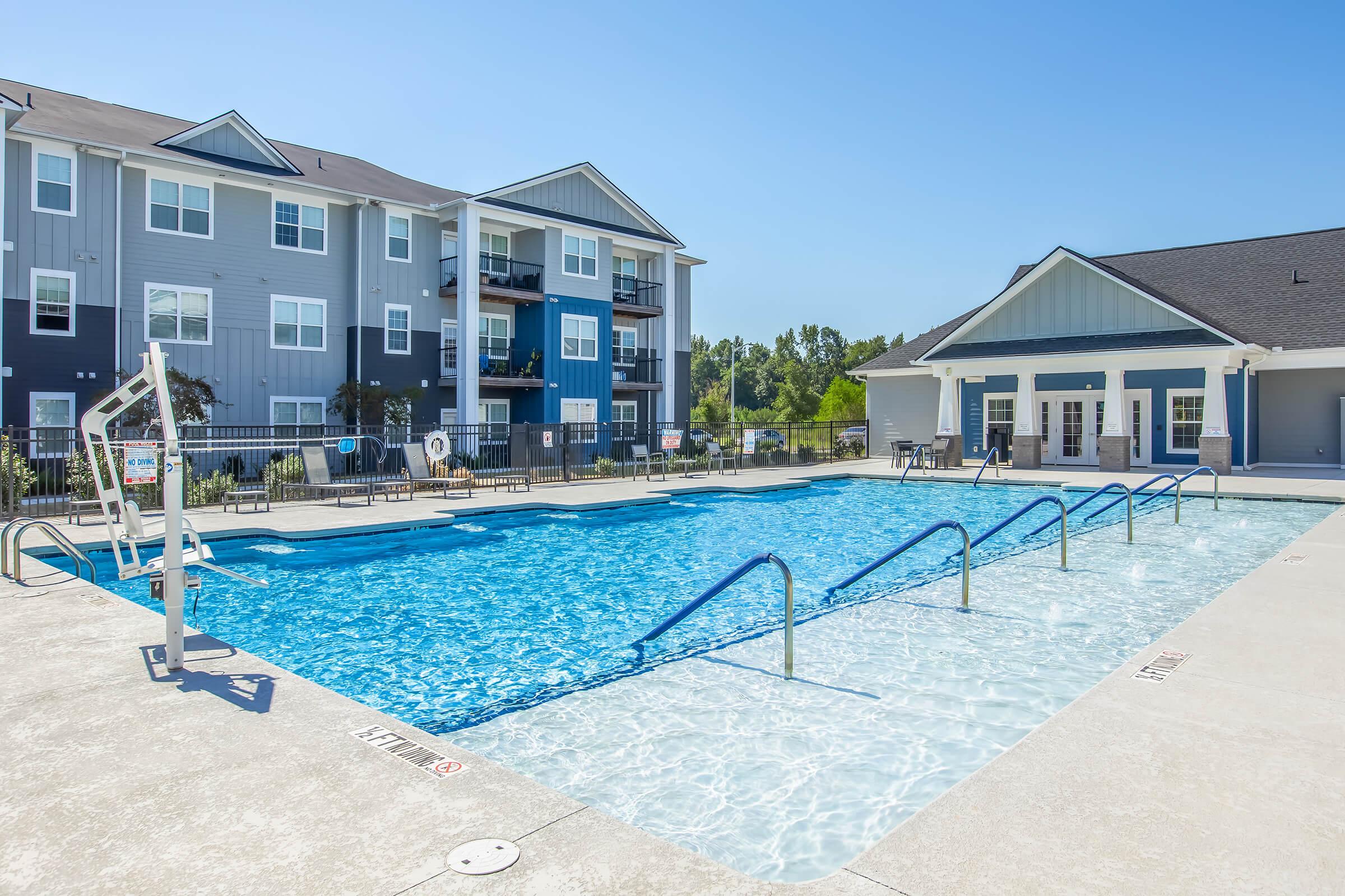 A clear blue swimming pool surrounded by lounge chairs and tables, with a backdrop of modern apartment buildings. The area is well-lit under a clear sky, creating a welcoming atmosphere for residents and guests. The pool features a shallow end and diving board.